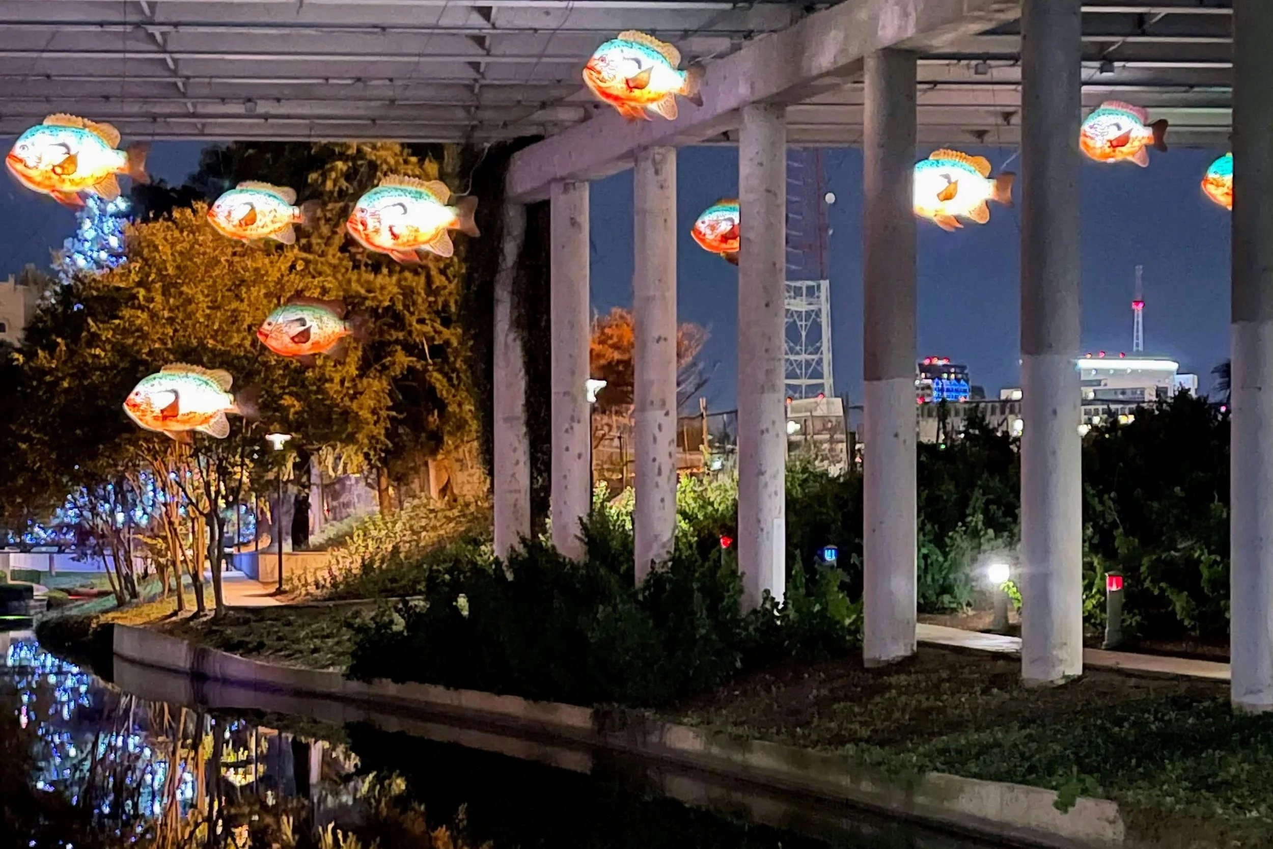 Several artistic fish hang from underneath a bridge with a river flowing underneath. Drives & Detours San Antonio River Walk hidden gems