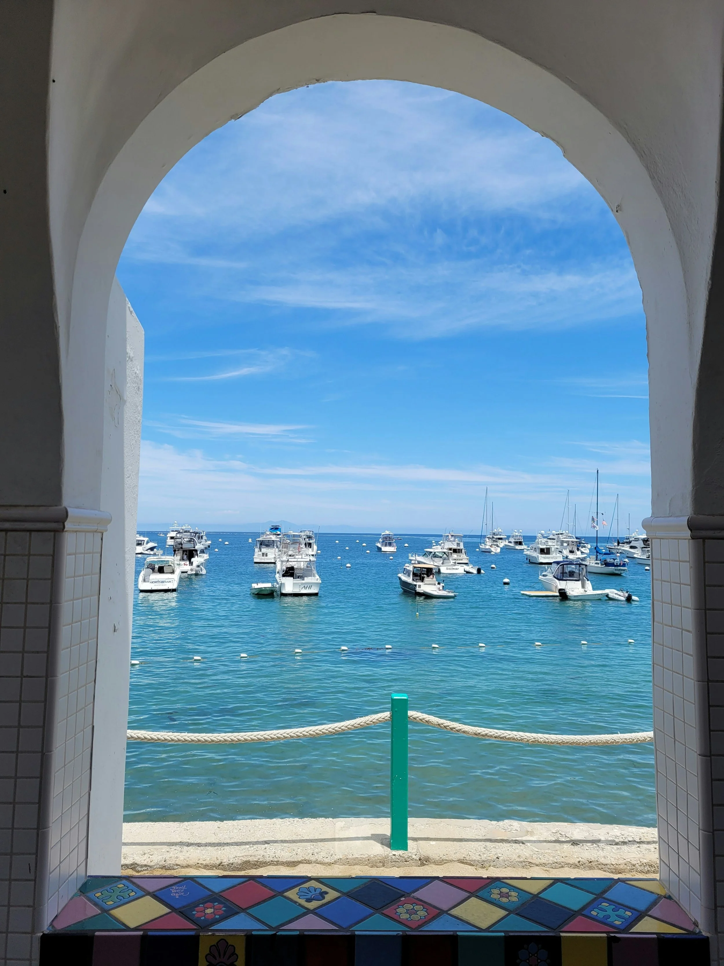 Small boats can be seen floating in Avalon harbor. The water is still. They are seen through a open, multi-colored tiled, window