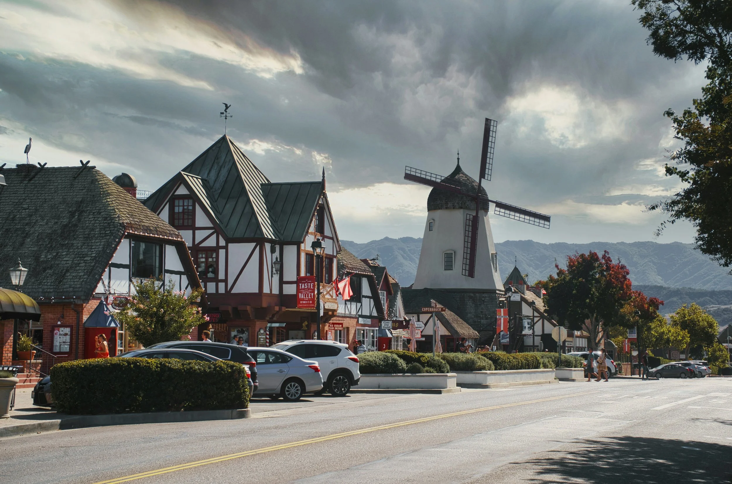 A street of old European wooden buildings. You can see the timber frames holding the roof up. At the end of the street is one of the large white windmills of Solvang, California
