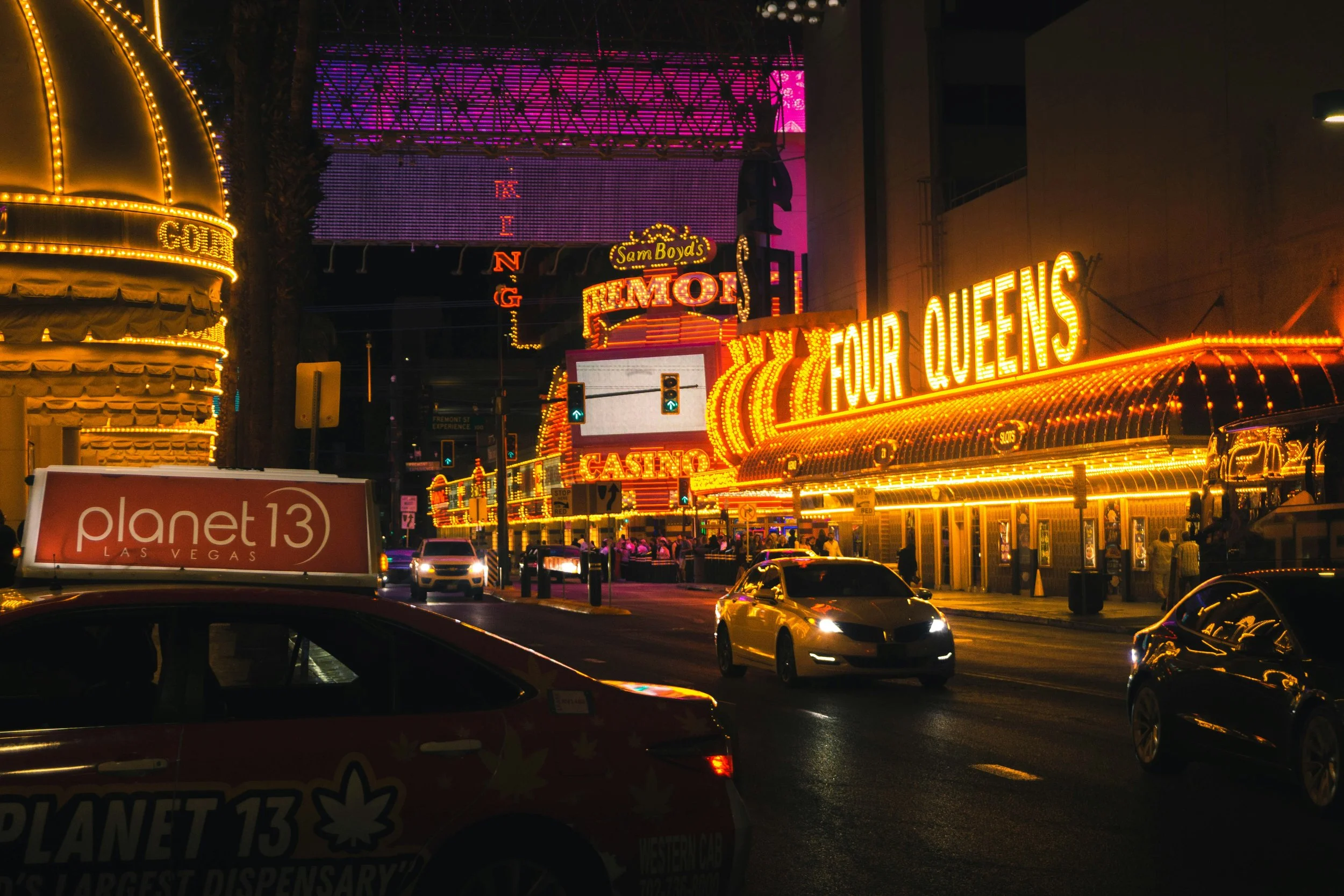 Cars drive past the Four Queens and Sam Boyd's Fremont Casinos at night. The casinos are lit up bright with flashing lights. Drives & Detours Las Vegas walking tour