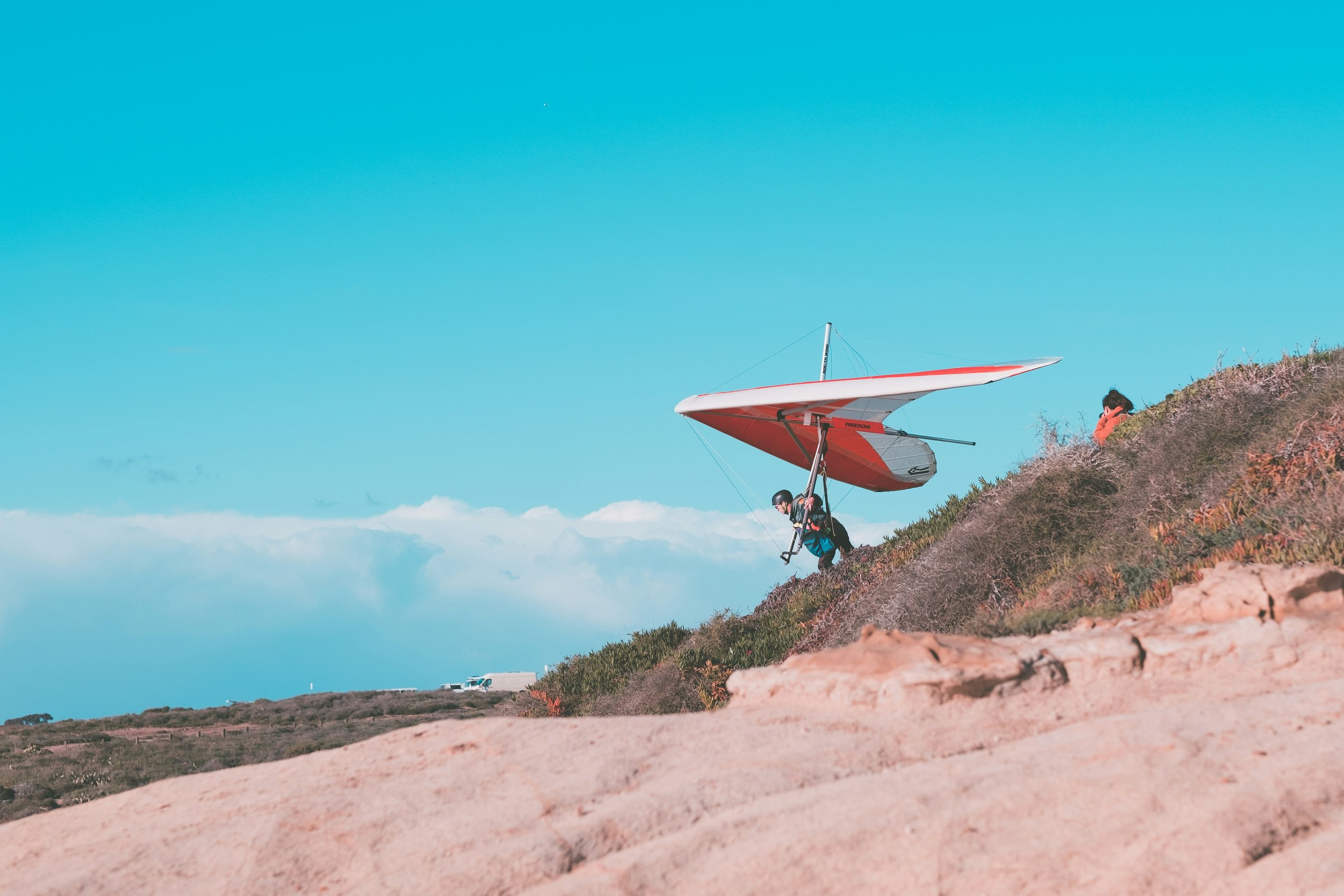 A person takes off on a red and white hang glider from a cliff. The sky is bright blue, with some clouds in the distance