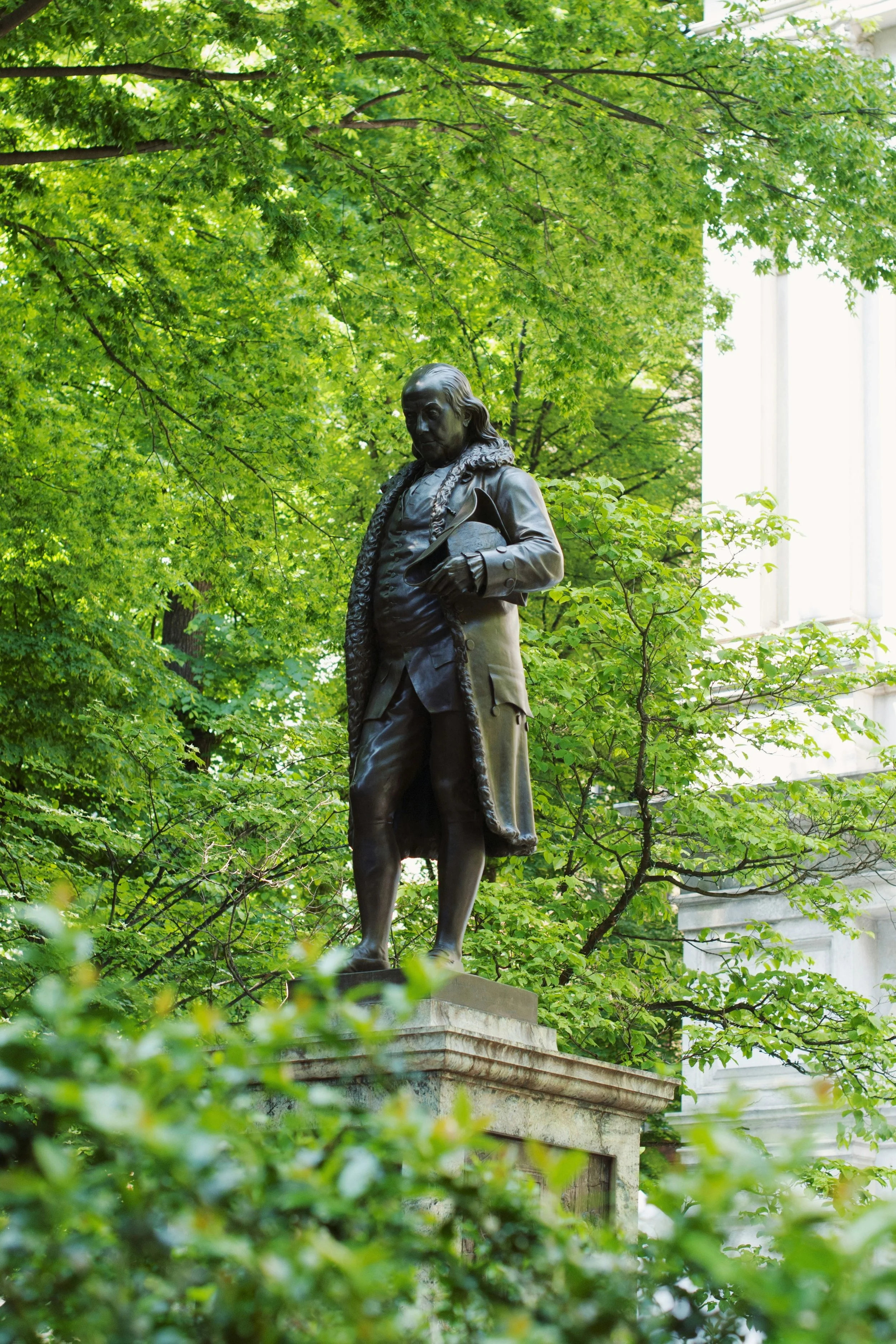 The statue of Benjamin Franklin located on the Freedom Trail in Boston is surrounded by green foliage. He looks quite portly and is looking down in thought