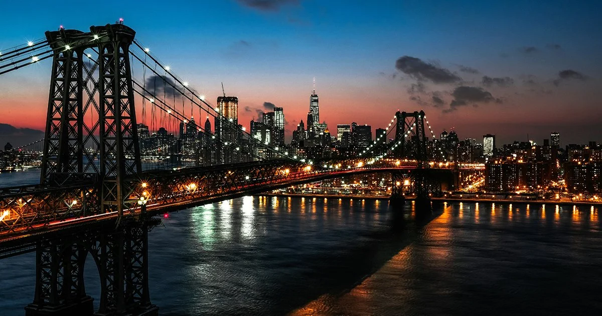 A bridge crosses the East River to Manhattan at dusk. New York City is lighting up under an orange sky