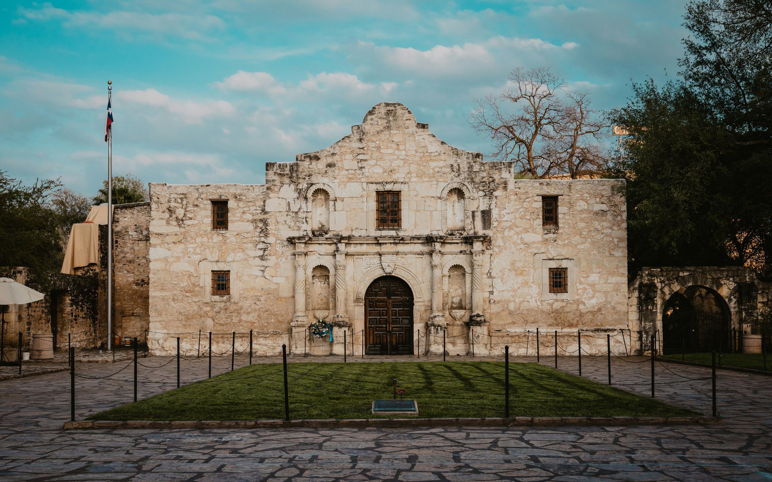 The ruins of the Alamo with a blue sky behind it. In front is a small park surrounded by old paving slabs, and a Texan Flag flies. Drives & Detours San Antonio self-guided tour