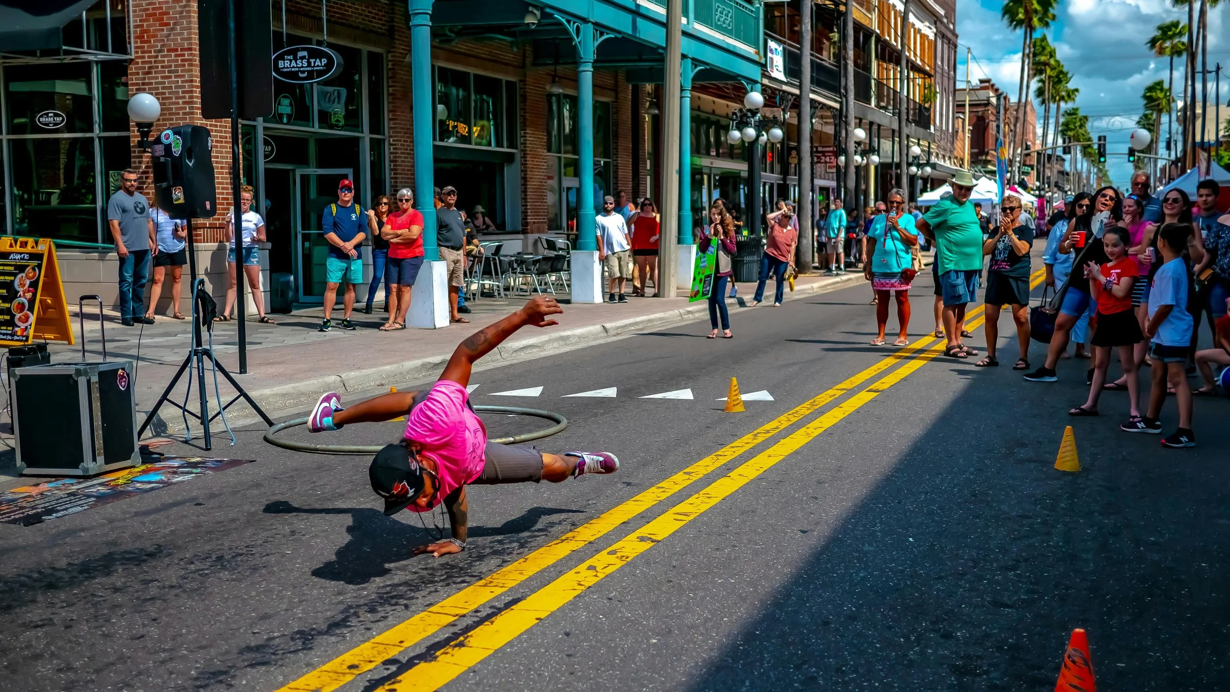 A man breakdances in Ybor City, Tampa. He's wearing a pink t-shirt and cap. A crowd watch and film on their phones