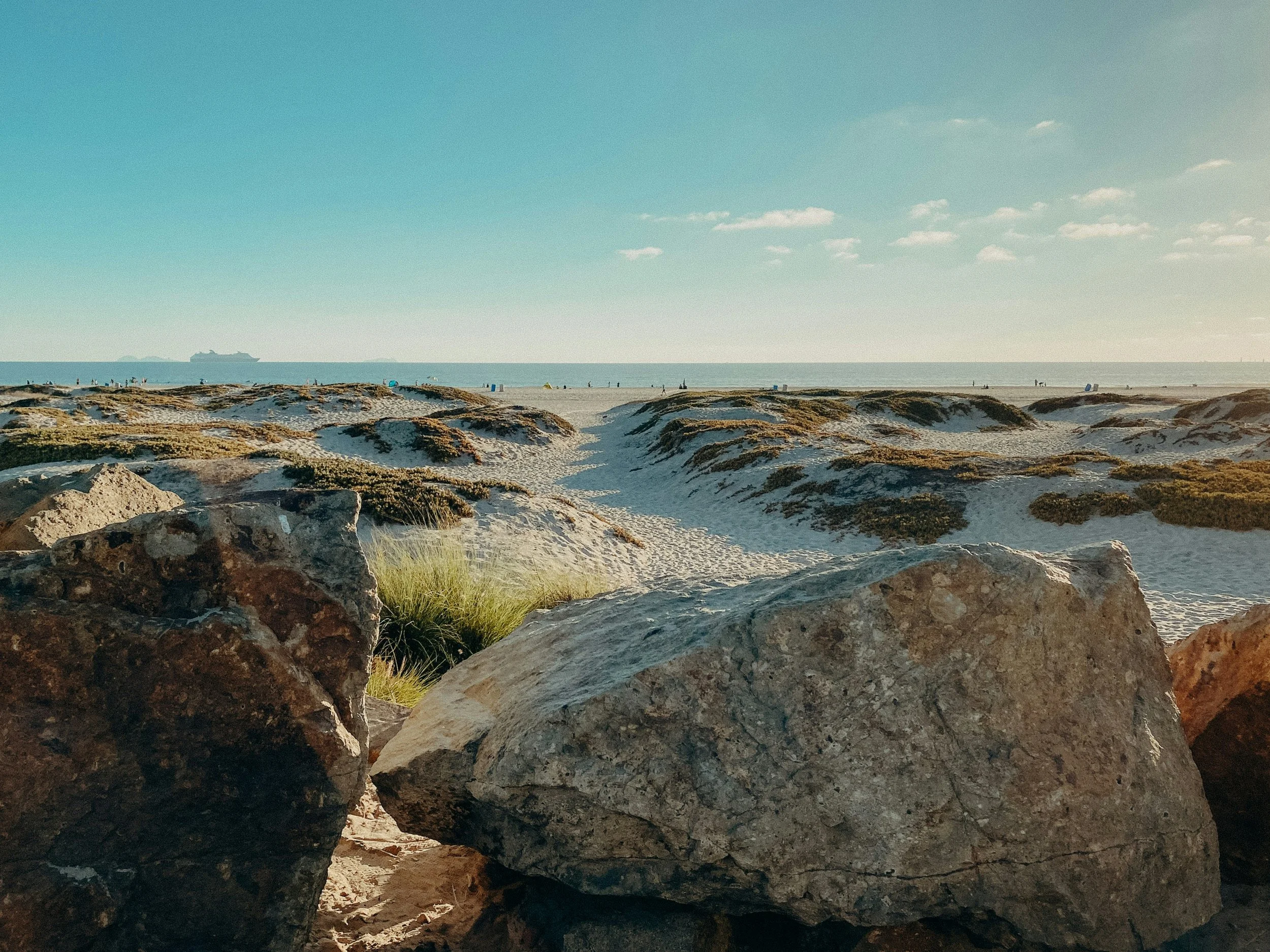 A rocky beach on Coronado Island. A cruise ship can be seen sailing on the horizon. A few cirrus clouds float in the sky