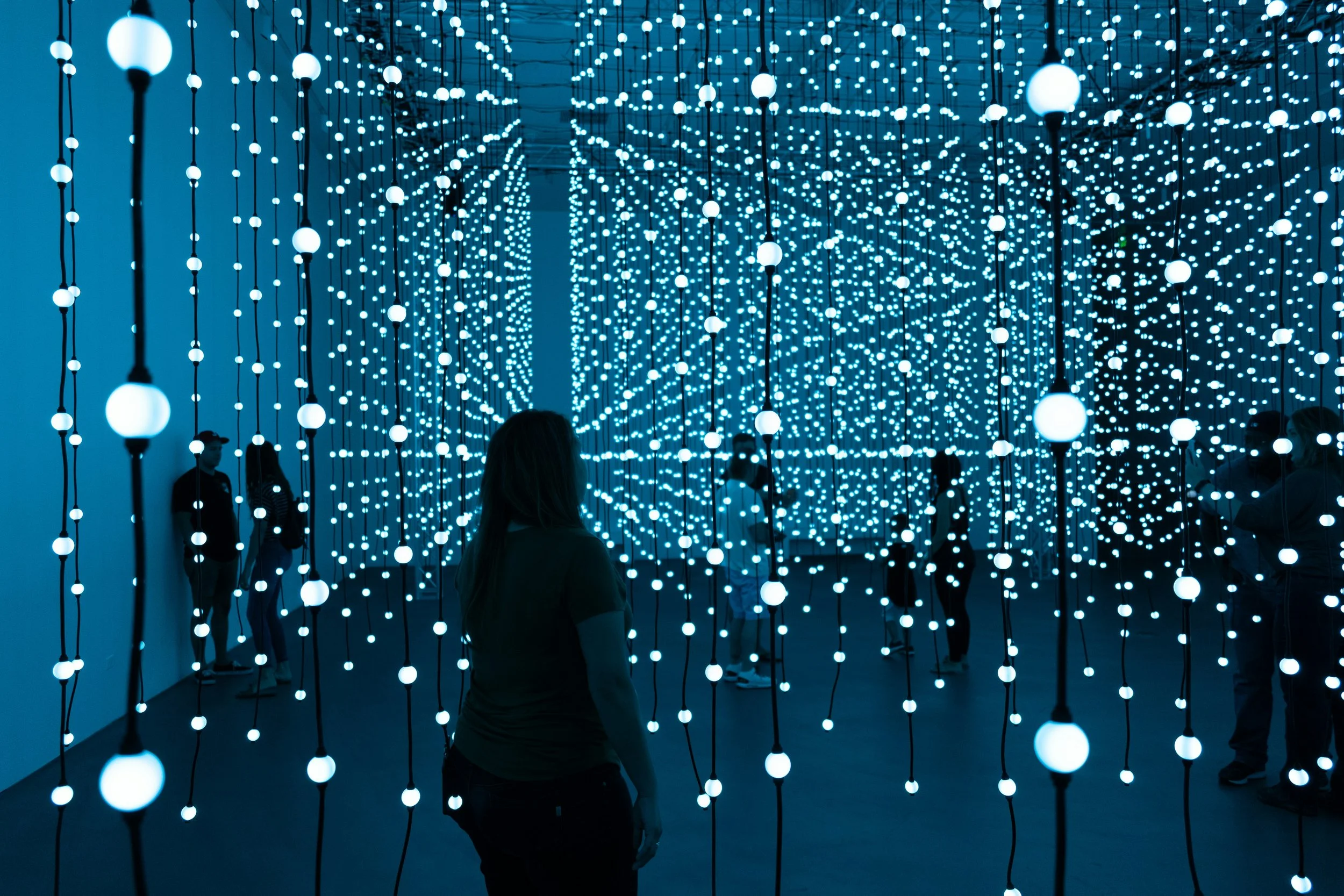 People walk through an art installation in Scottsdale, Arizona. Thousands of dim lights hang from a ceiling, and the room is a blue colour. The people are silhouettes. Drives & Detours Scottsdale self-guided tour