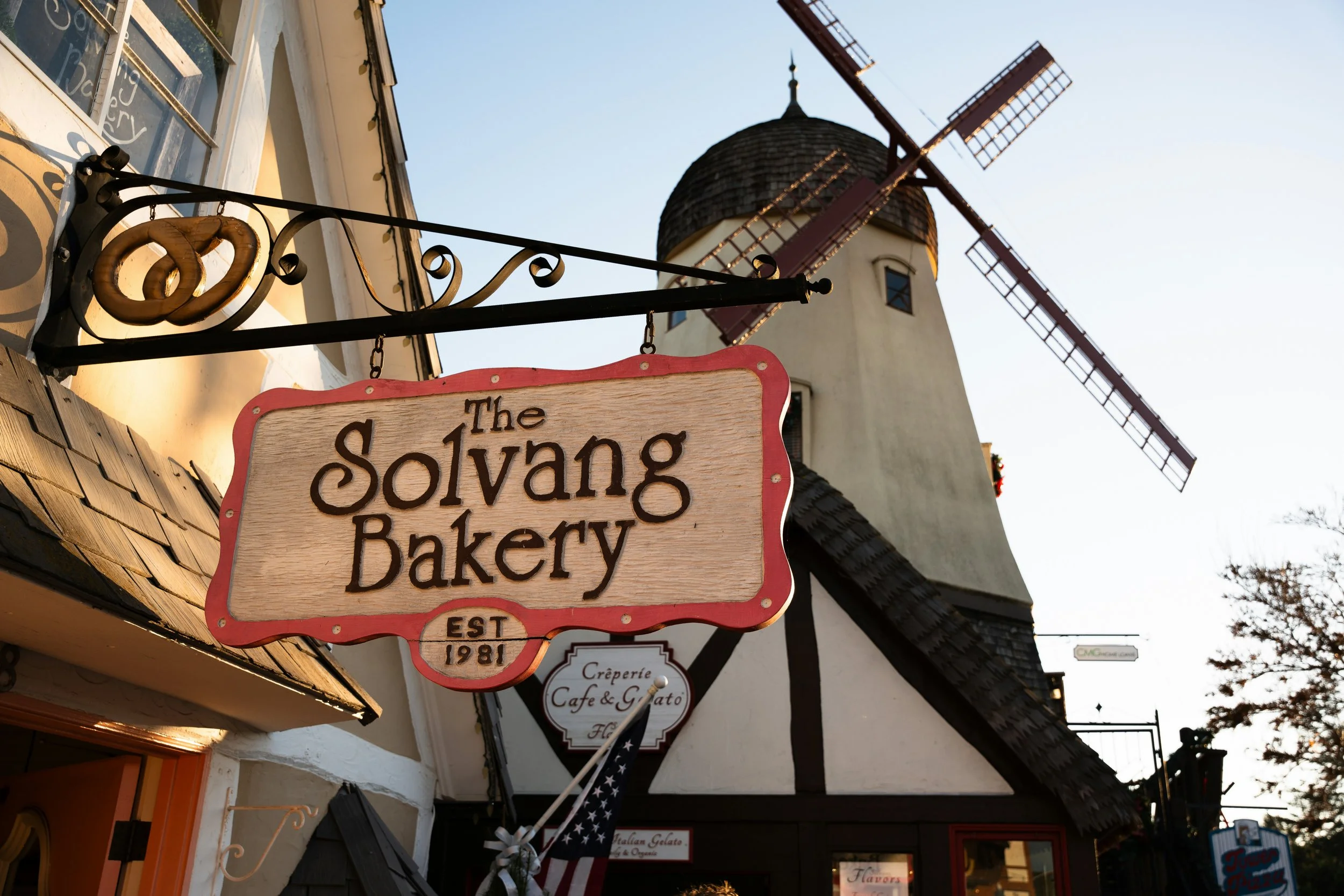 A wooden sign hanging from a shopfront reads: The Solvang Bakery, Est 1981. Behind it is a large white windmill, in the classic style, with four sails
