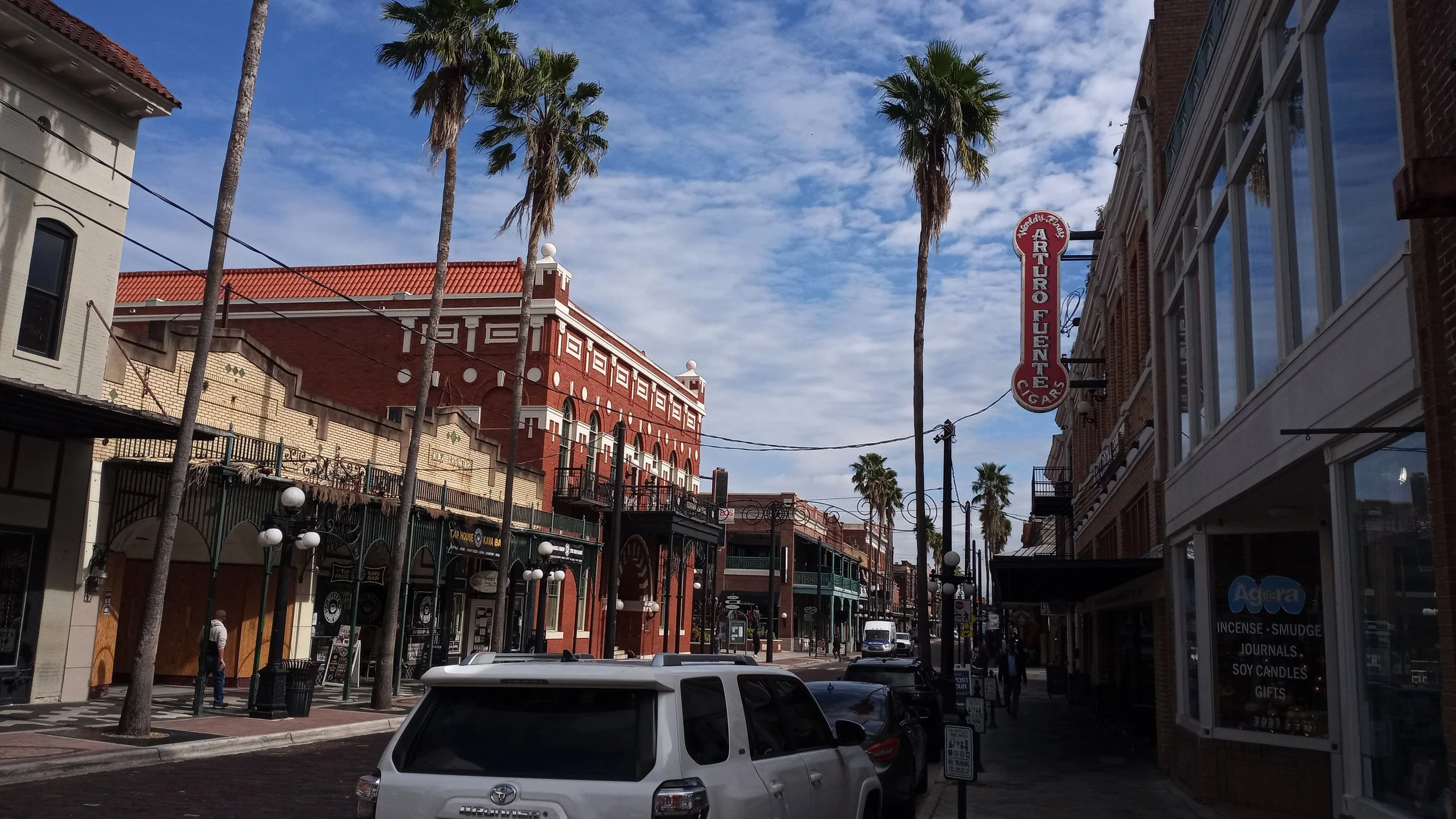 Cars are parked along 7th Avenue in Ybor City, Tampa. There is a large brick building further down the street, and tall palm trees