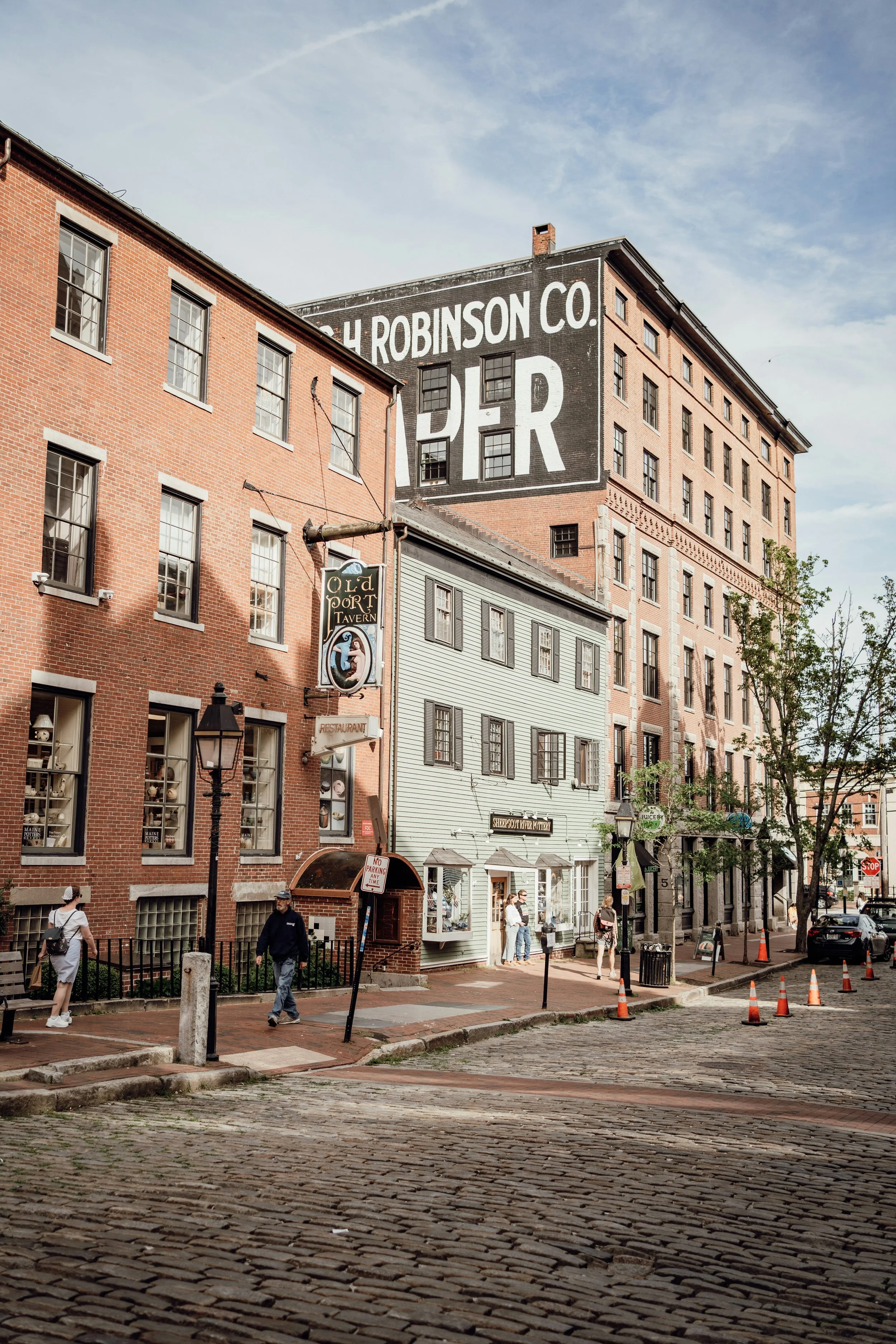 People walk along a cobbled street in Portland, Maine. A wooden sign hangs from one of the buildings, reading,Old Port Tavern, while another is painted on a brick building, but its unreadable