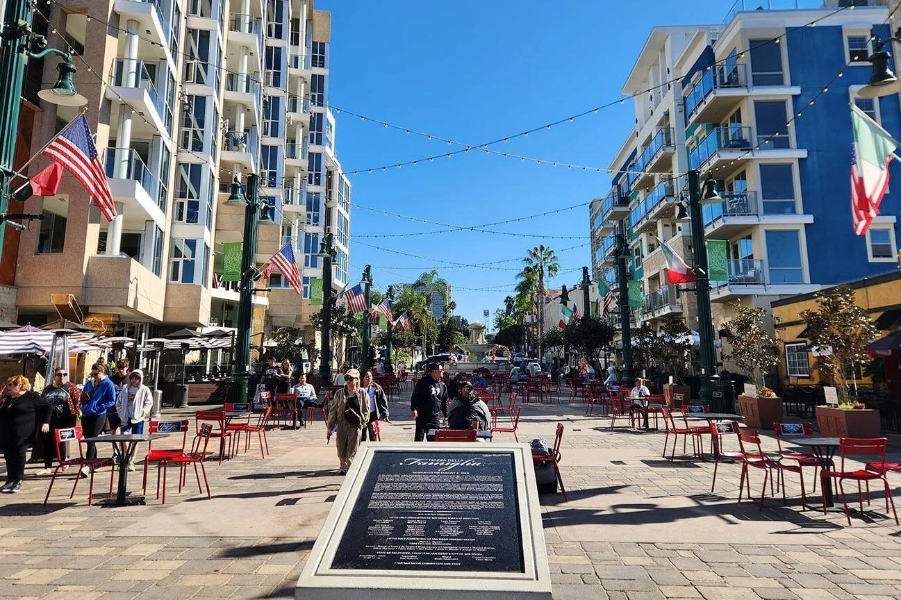 The large Piazza della Famiglia in San Diego. Italian and American flags from the buildings at its edge, and the square is full of coffee tables and people milling around in the sun.