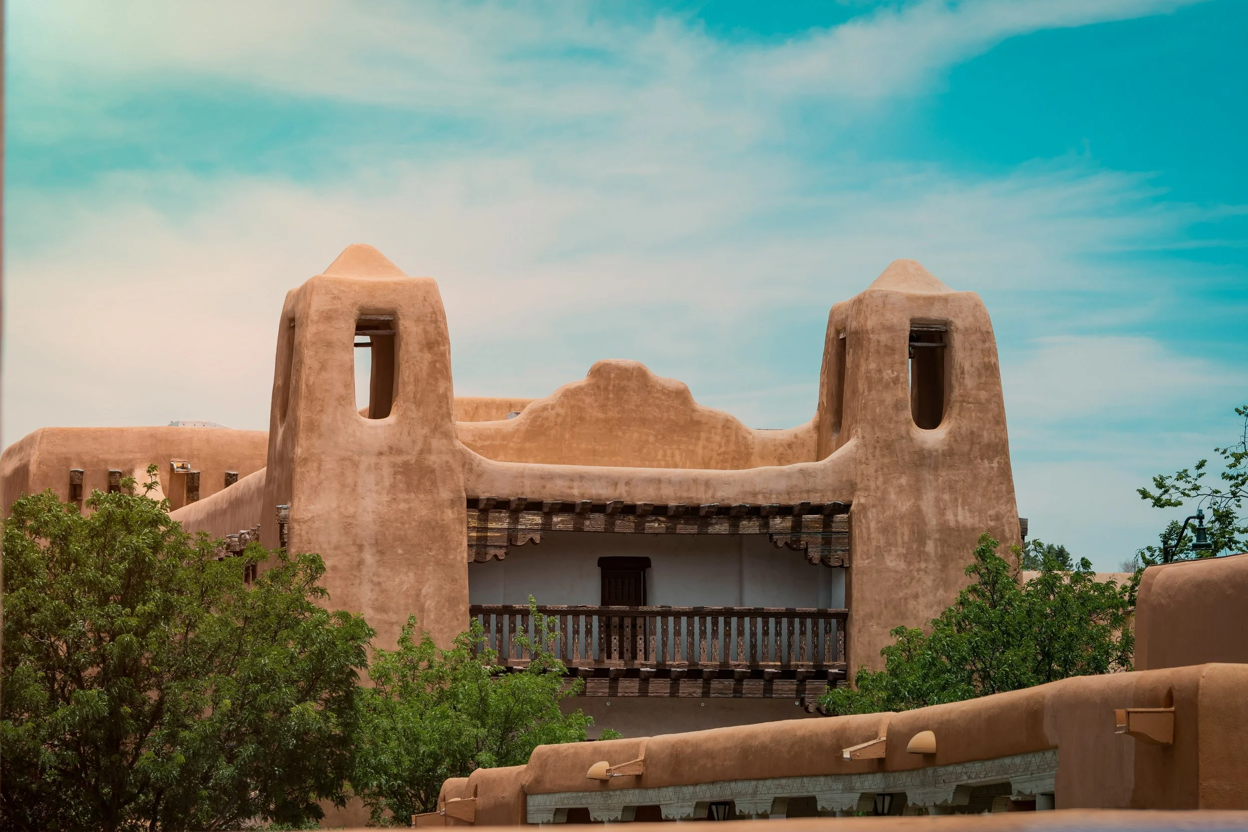 An adobe building in Santa Fe with a balcony. It is surrounded by blue sky and some trees