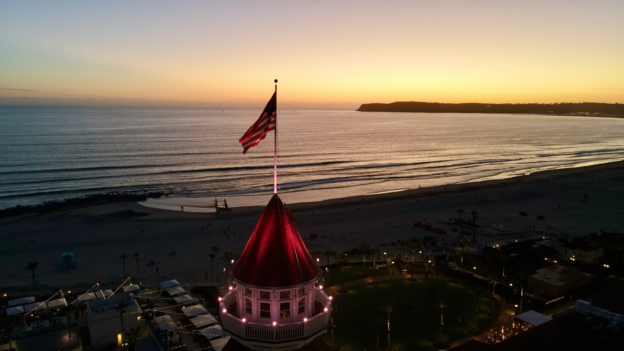 An American flag flies from the roof of the Hotel Del Coronado at sunset. Lights are beginning to come on around the hotel, but the beach is still dark. The sky is a bright orange