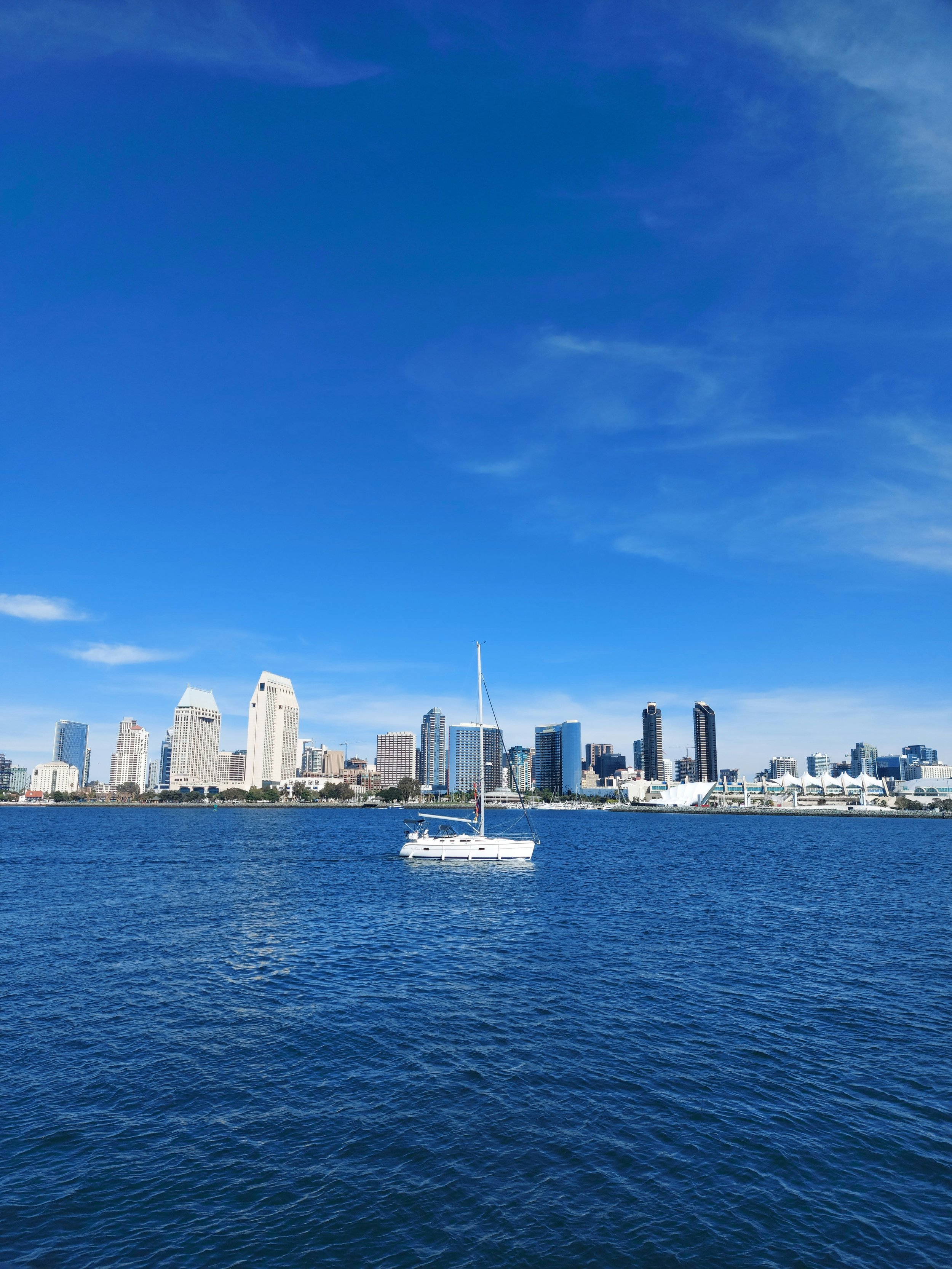 A small, white yacht floats in the bay between Coronado Island and San Diego. The skyline is bathed in sunlight and the sky is bright blue