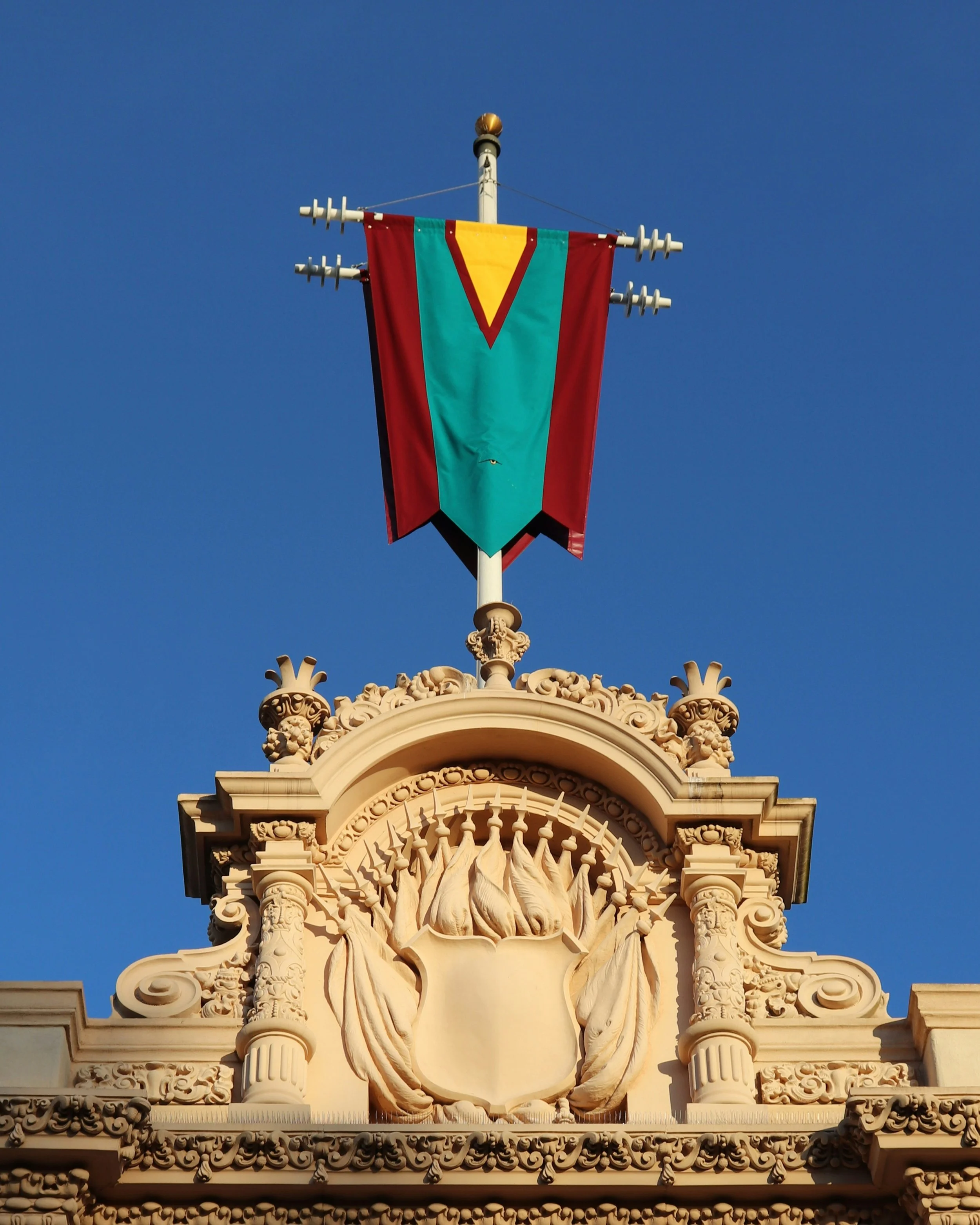 A yellow, teal and deep red flag hangs on top of a building in Balboa Park, San Diego. Underneath, a sculture in the stone of the building shows a shield surrounded by flags