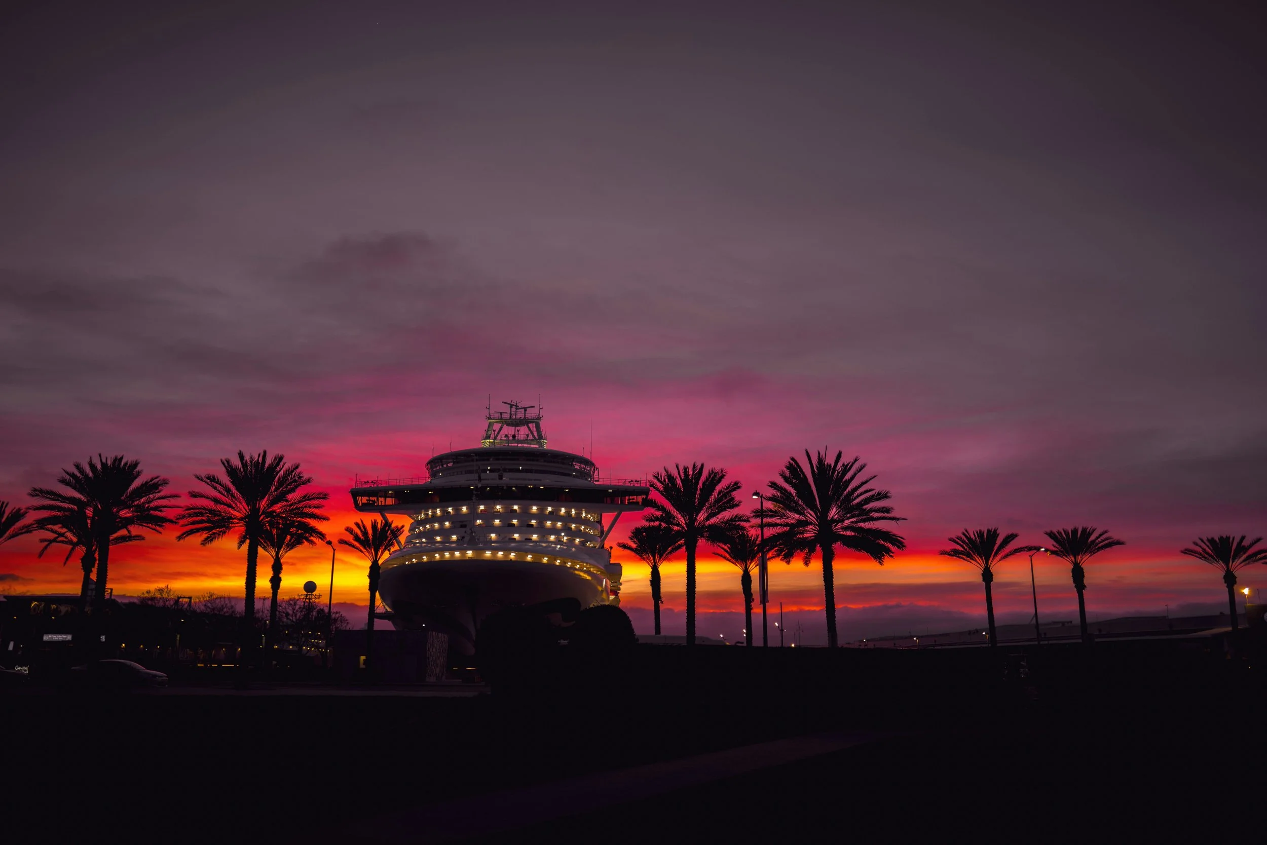 A cruise ship is docked at the San Diego Embarcadero at sunset. The sky is a vivid purple and orange. The ship and the palm trees that surround it are silhouettes
