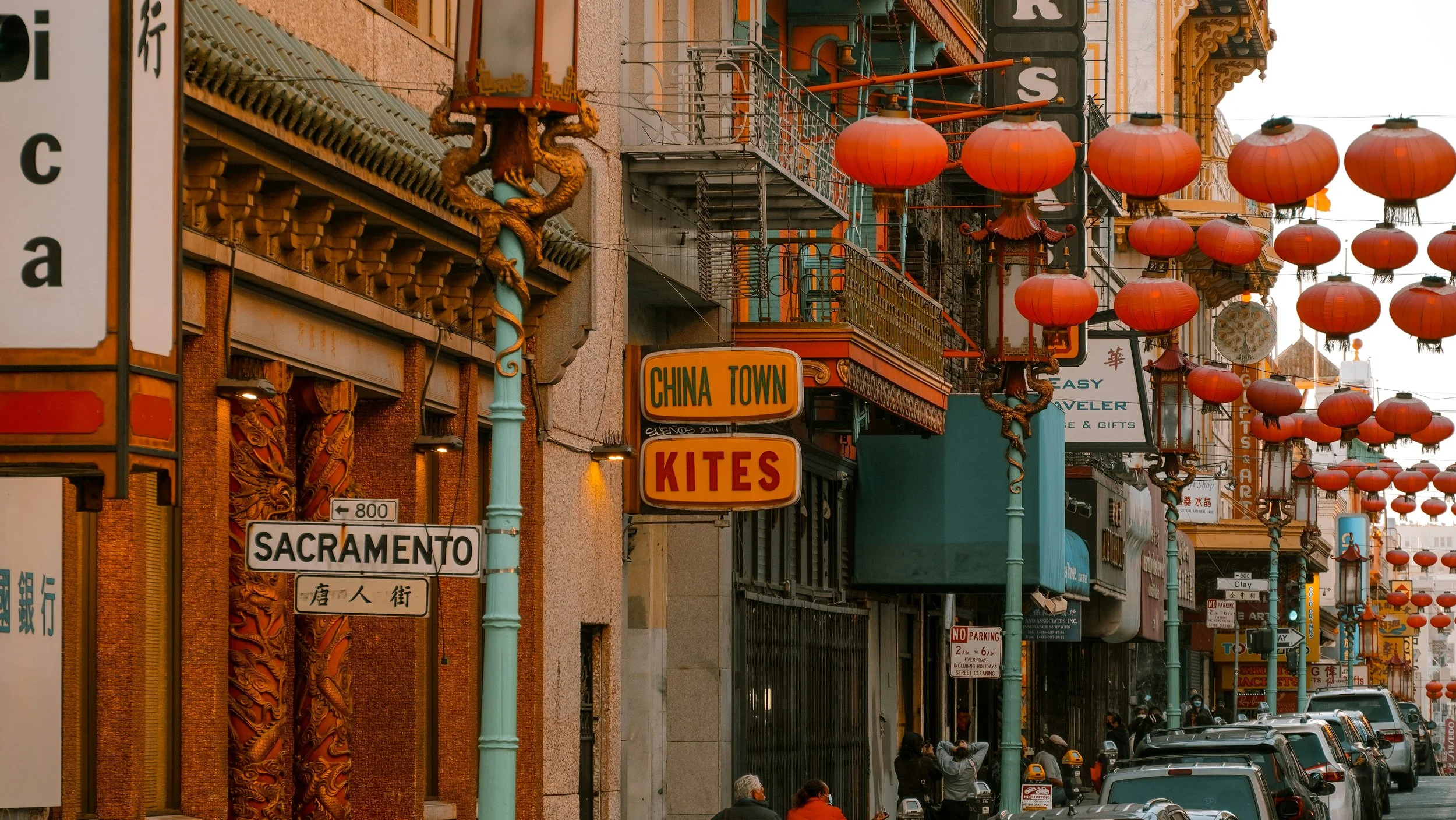 A streetscape in San Francisco Chinatown. Lanterns hang across the street, a street sign reads "Sacramento", and a sign outside a shop reads: "China Town Kites." Dragons decorate the lampposts