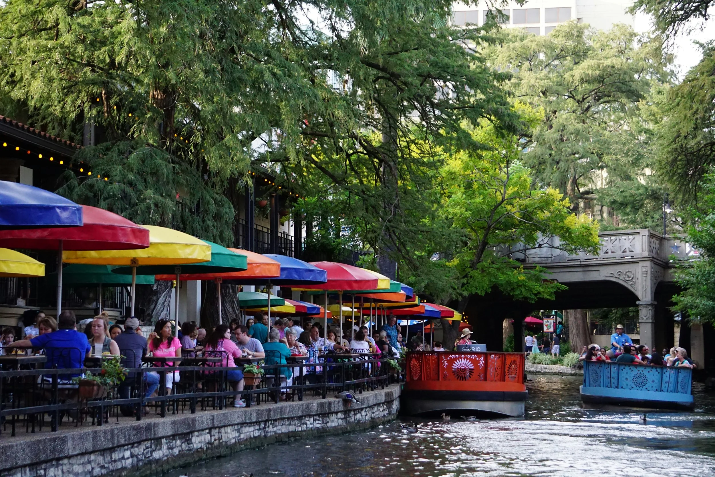 Red and blue pleasure boats sail past a restaurant with multi-colored umbrellas on the San Antonio River Walk