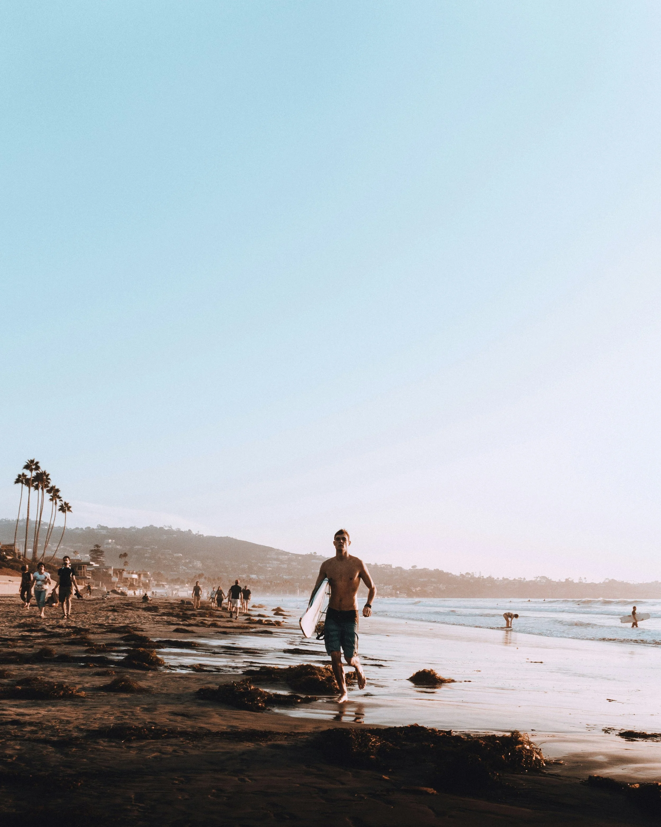 A surfer runs along the beach at La Jolla. He is topless, in shorts and carrying a surf board. The beach is full of people and the sky is a bright blue