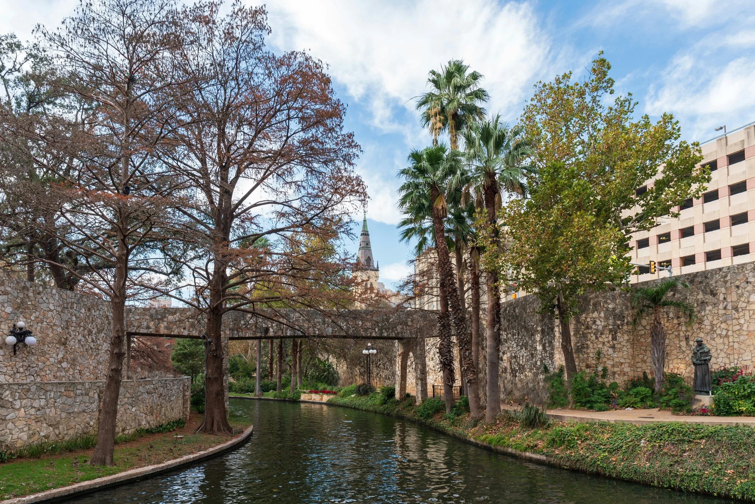 Palm trees line a river covered with a stone bridge and lined with paths. An old statue of a priest is on the banks. Drives & Detours Best places to eat in San Antonio