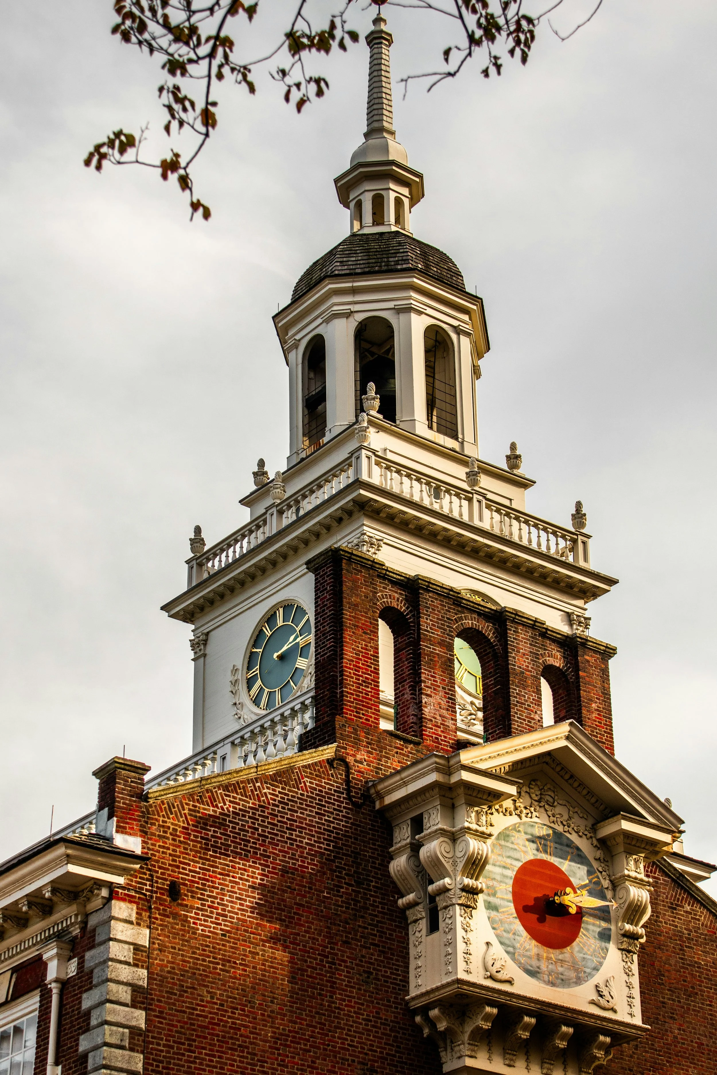 A detail of the clock tower on Independence Hall in Philadelphia. One clock has a red centre, with Roman numerals written in gold on a blue background. The other on a different building has a blue background with the Roman numerals on a metal frame