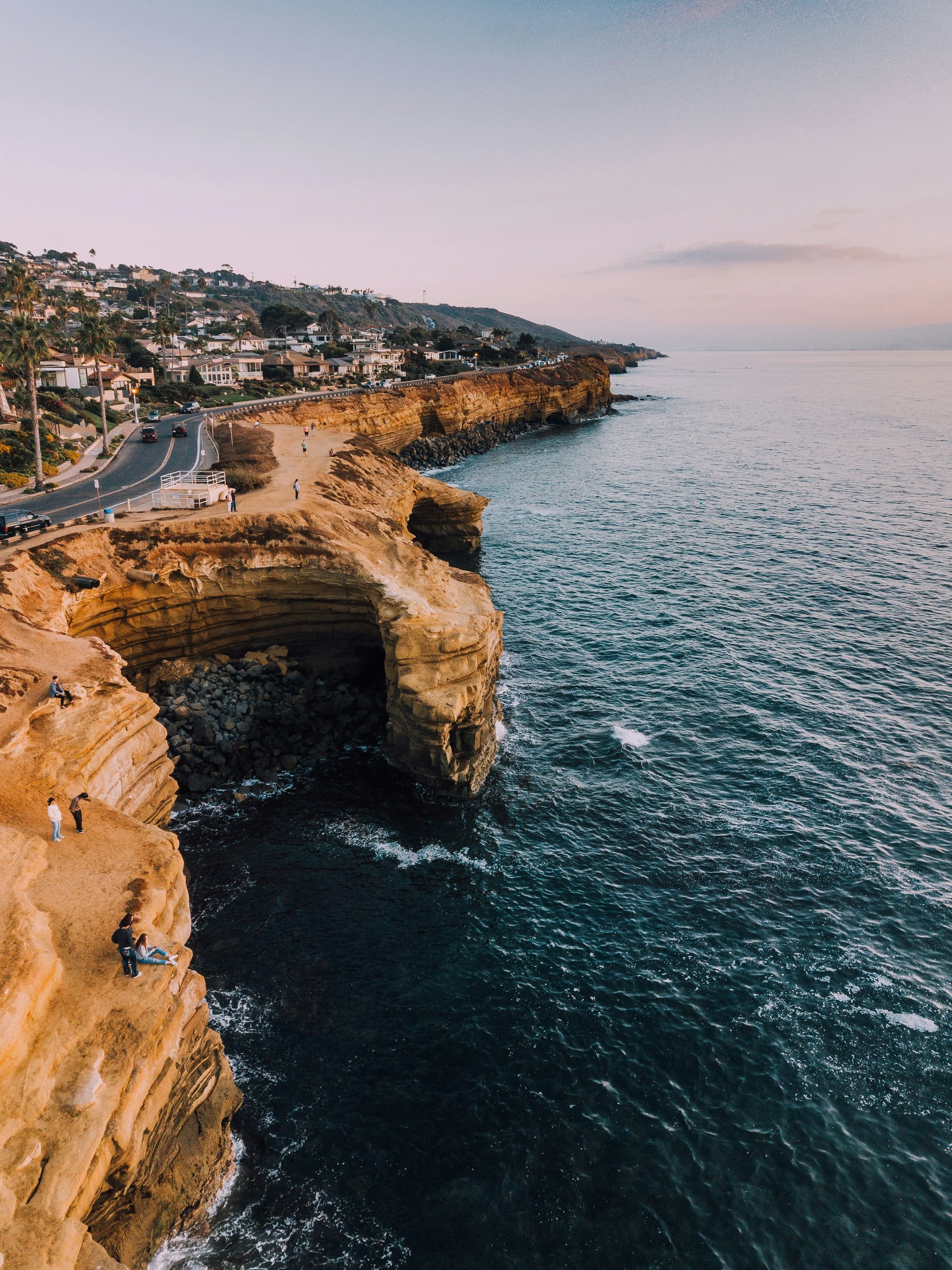 A road winds its way along a tall cliff in San Diego. The cliff is orange roack, and drops down to the still dark blue water of the Pacific