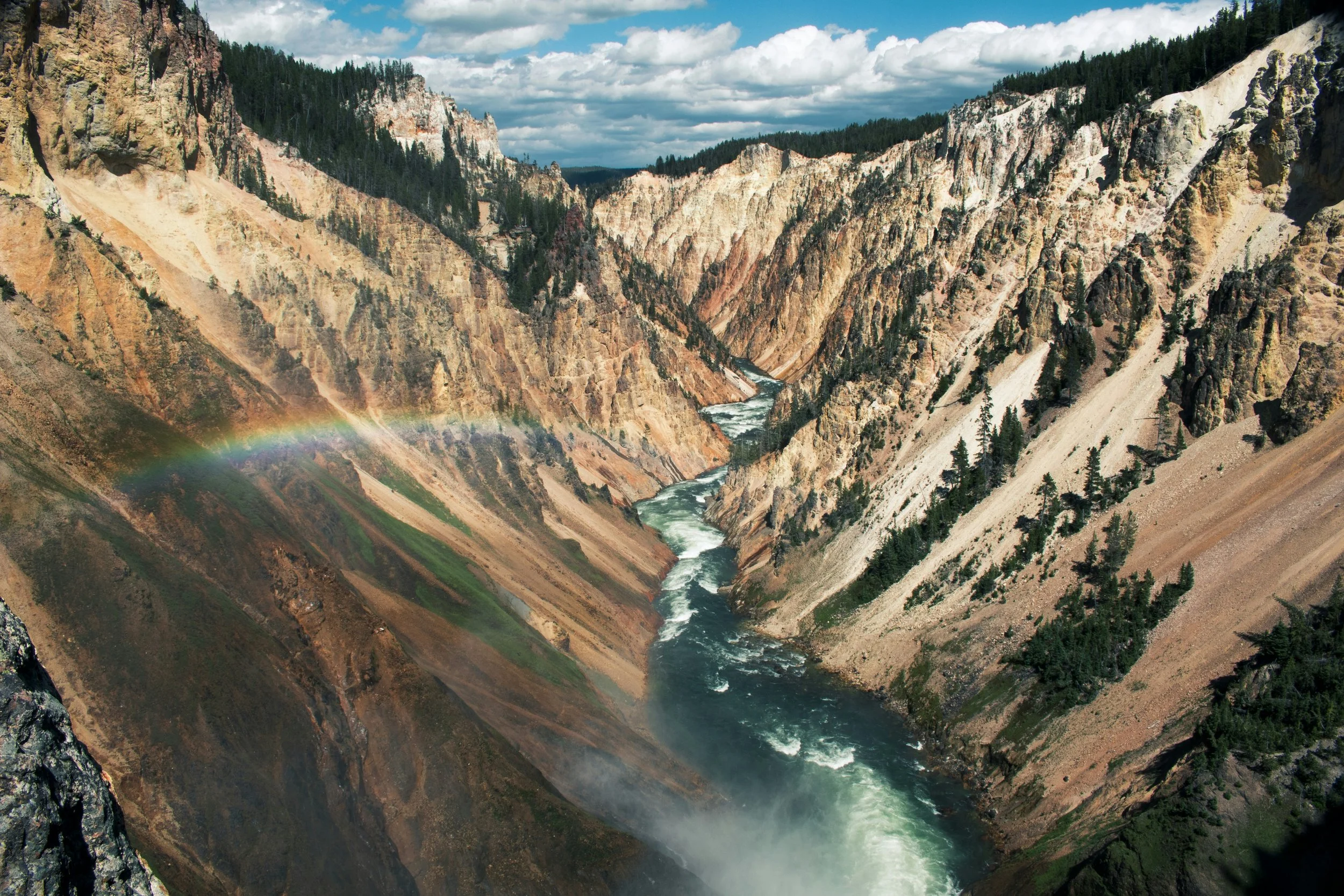 A rainbow arcs over the Grand Canyon of the Yellowstone. A river pours through the steep valley. Drives & Detours Yellowstone driving tour