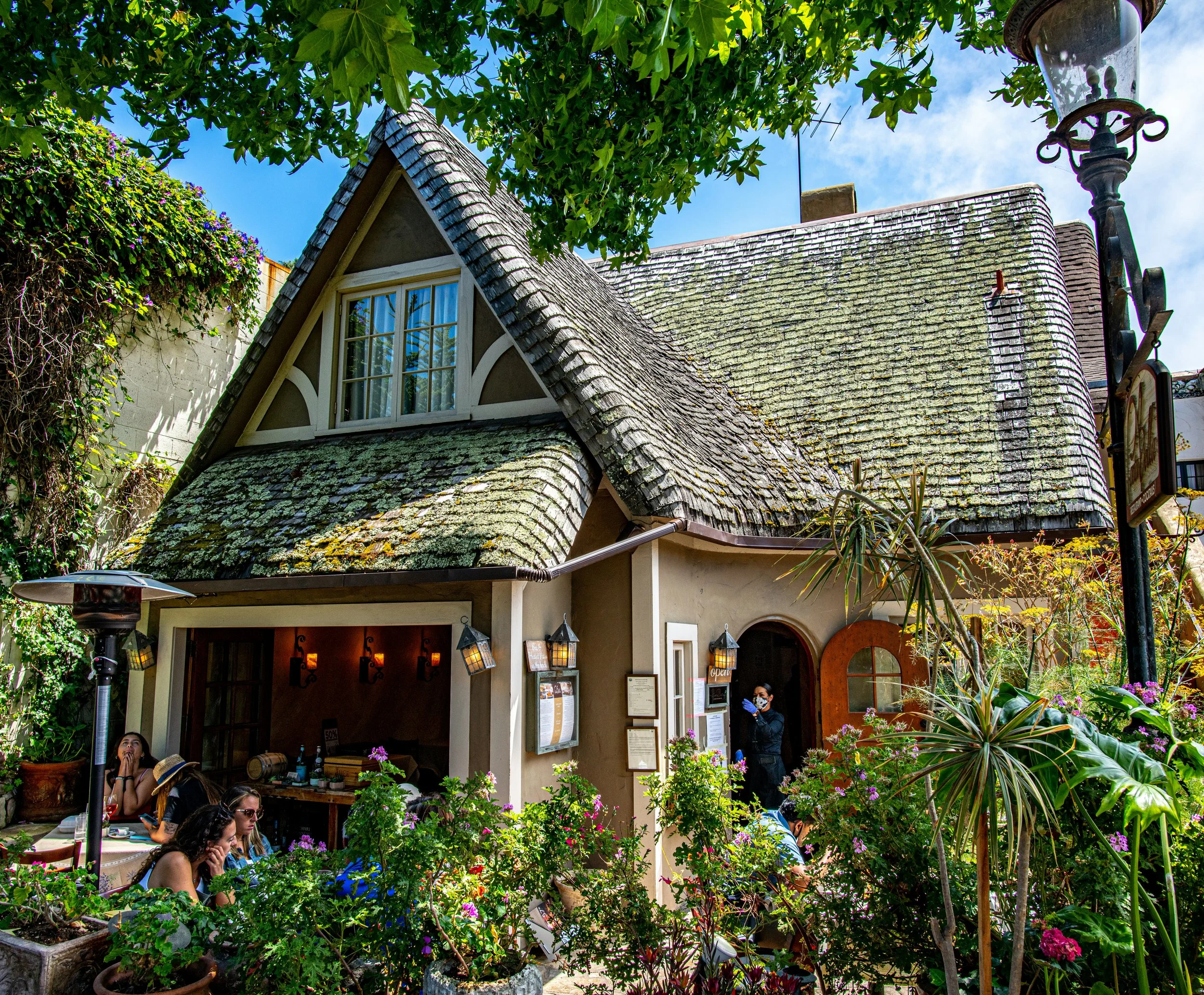 People drink coffee outside a cottage in Carmel-by-the-Sea, that has a Old World style of hanging lanterns and gabled eaves