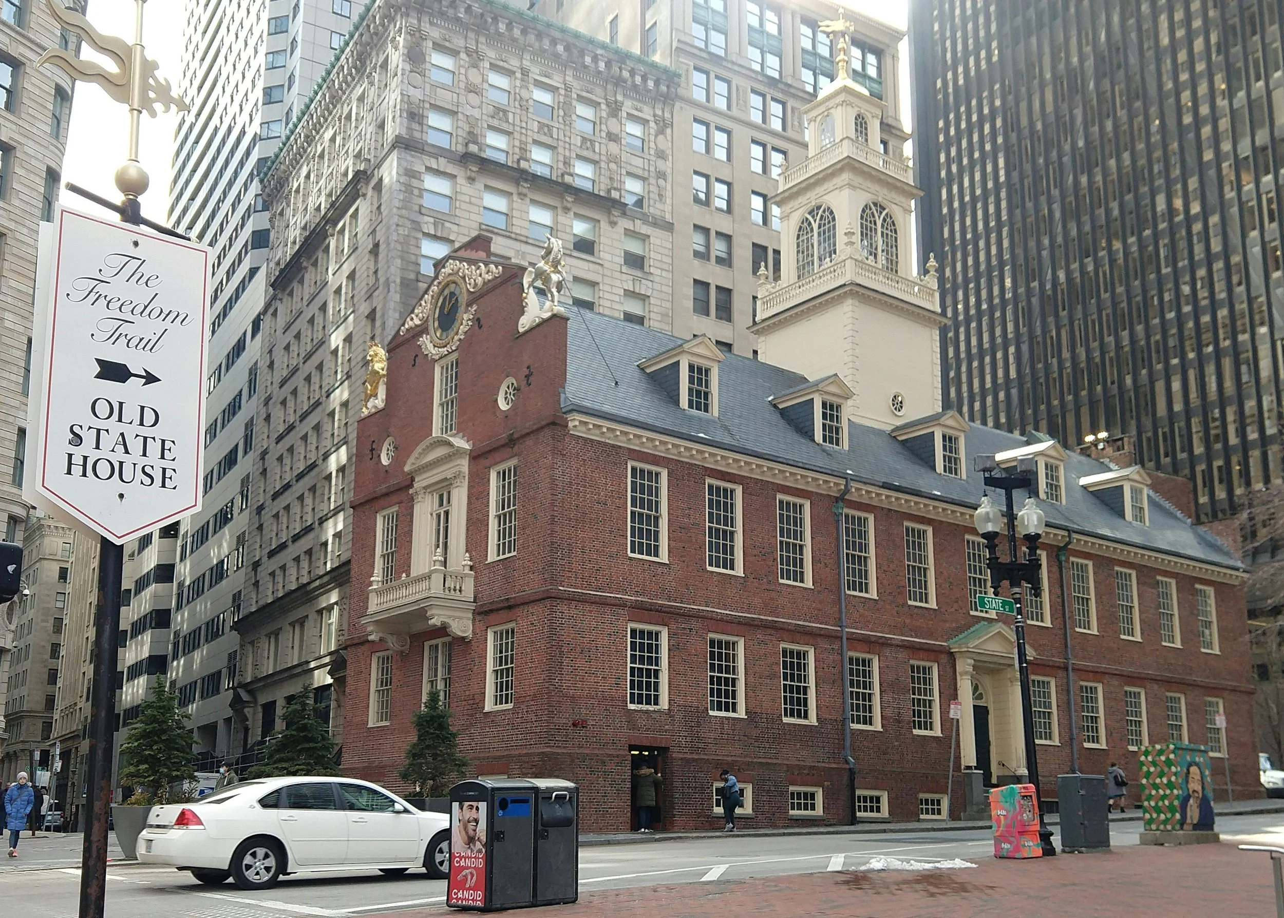 The Old State House in Boston is surrounded by skyscrapers. The three-storey, brick building is tiny in comparison. It has a white tower on top. A sign points to it, reading: The Freedom Trail, Old State House