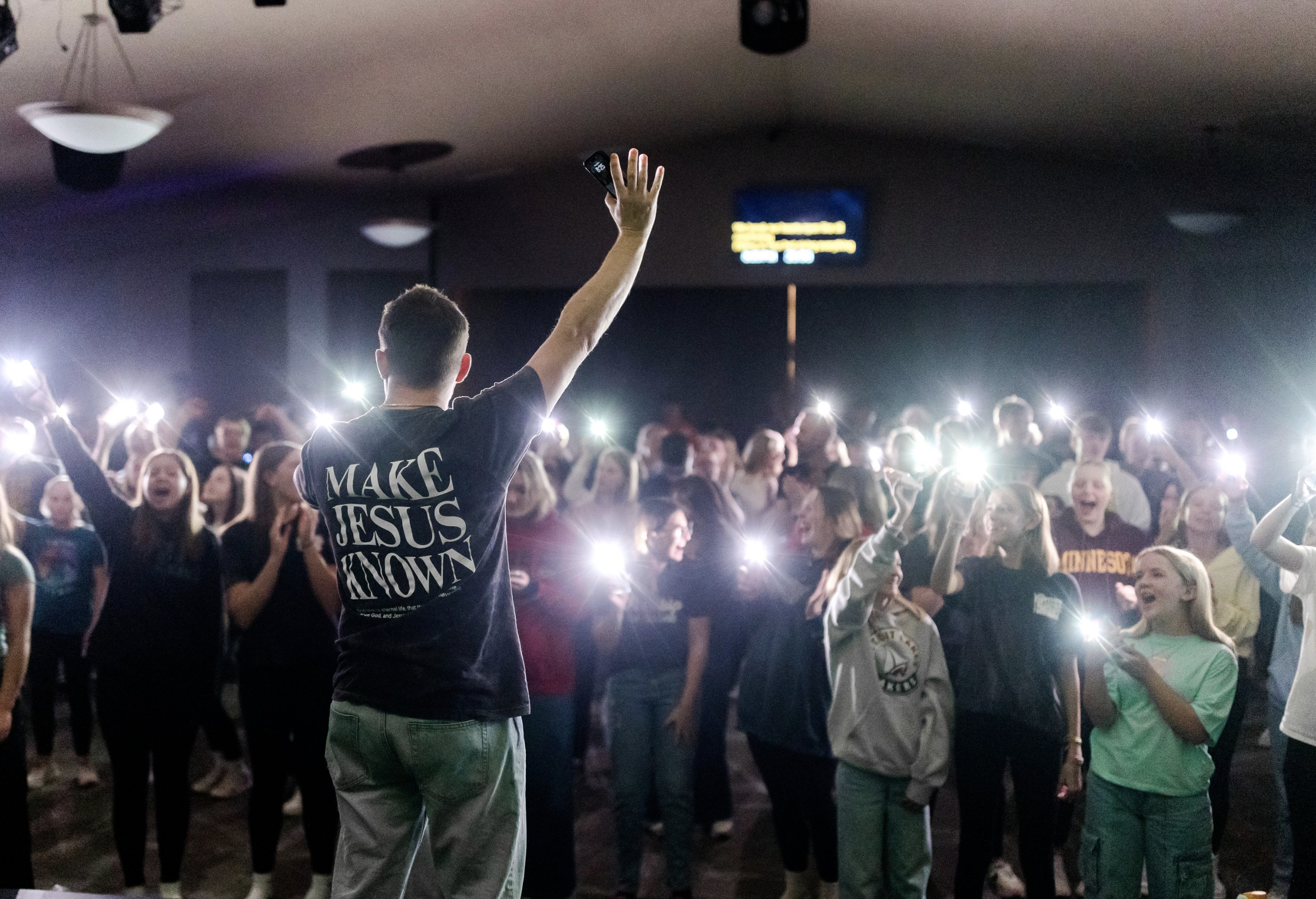 A man standing on a stage with his back to the camera, wearing a shirt that says 'Make Jesus Known,' addressing a large crowd of young people in an indoor setting. The audience is smiling, cheering, and holding up their phones with lights on.
