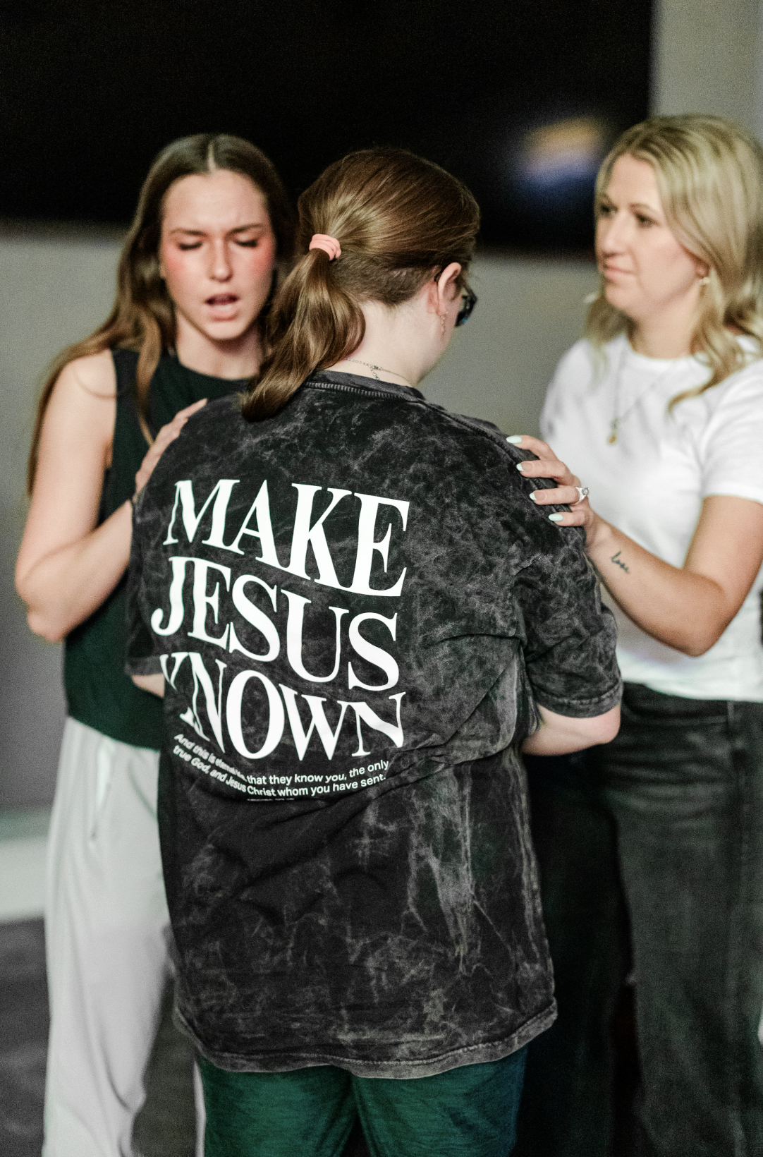 Three women gathered closely, one with her back to the camera wearing a black shirt with 'MAKE JESUS KNOWN' printed on it. The other two women face her, both appearing emotional, with one gently holding her shoulders.