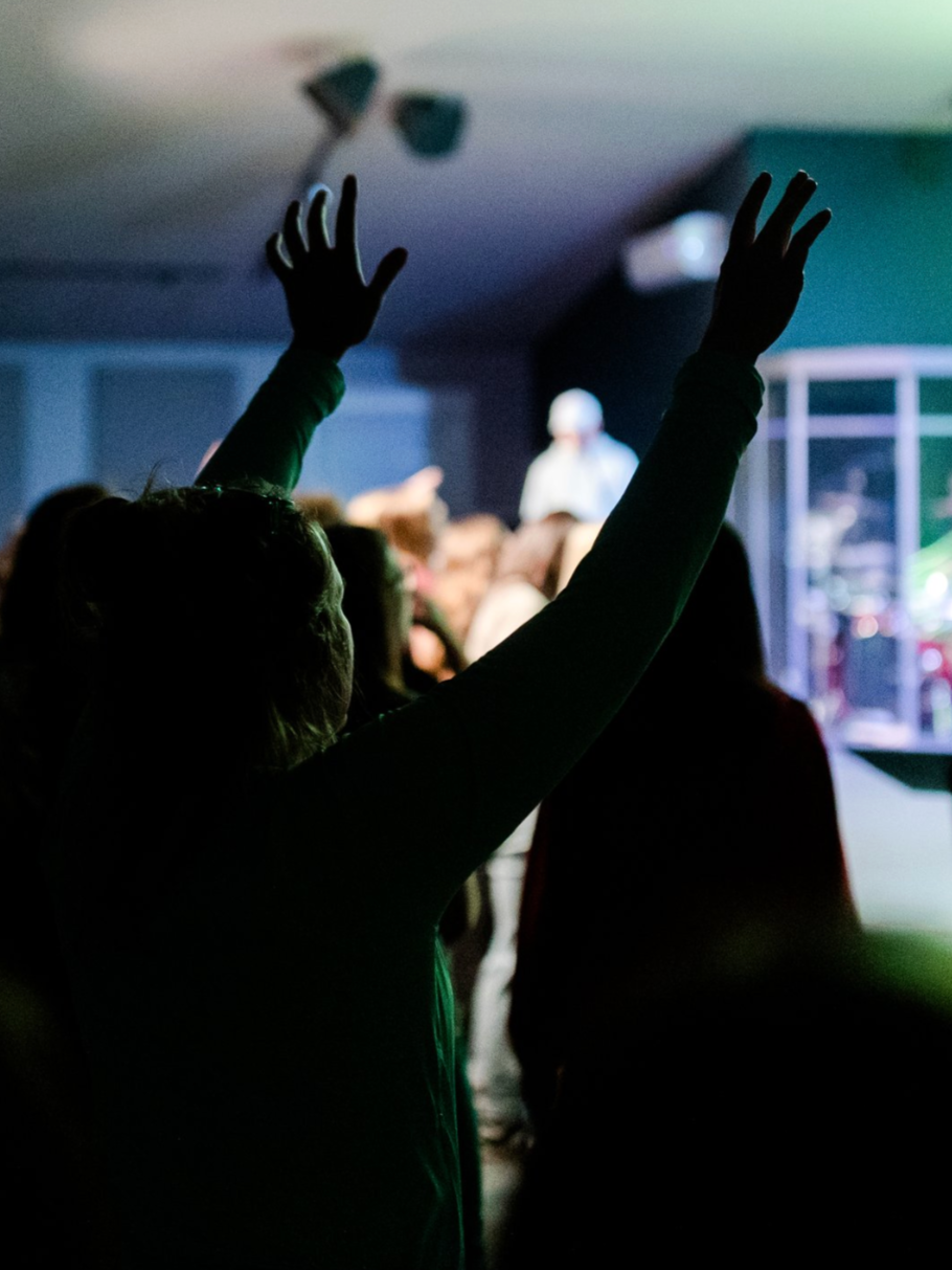 Person raising hands during a concert or worship event with a blurred stage in the background.