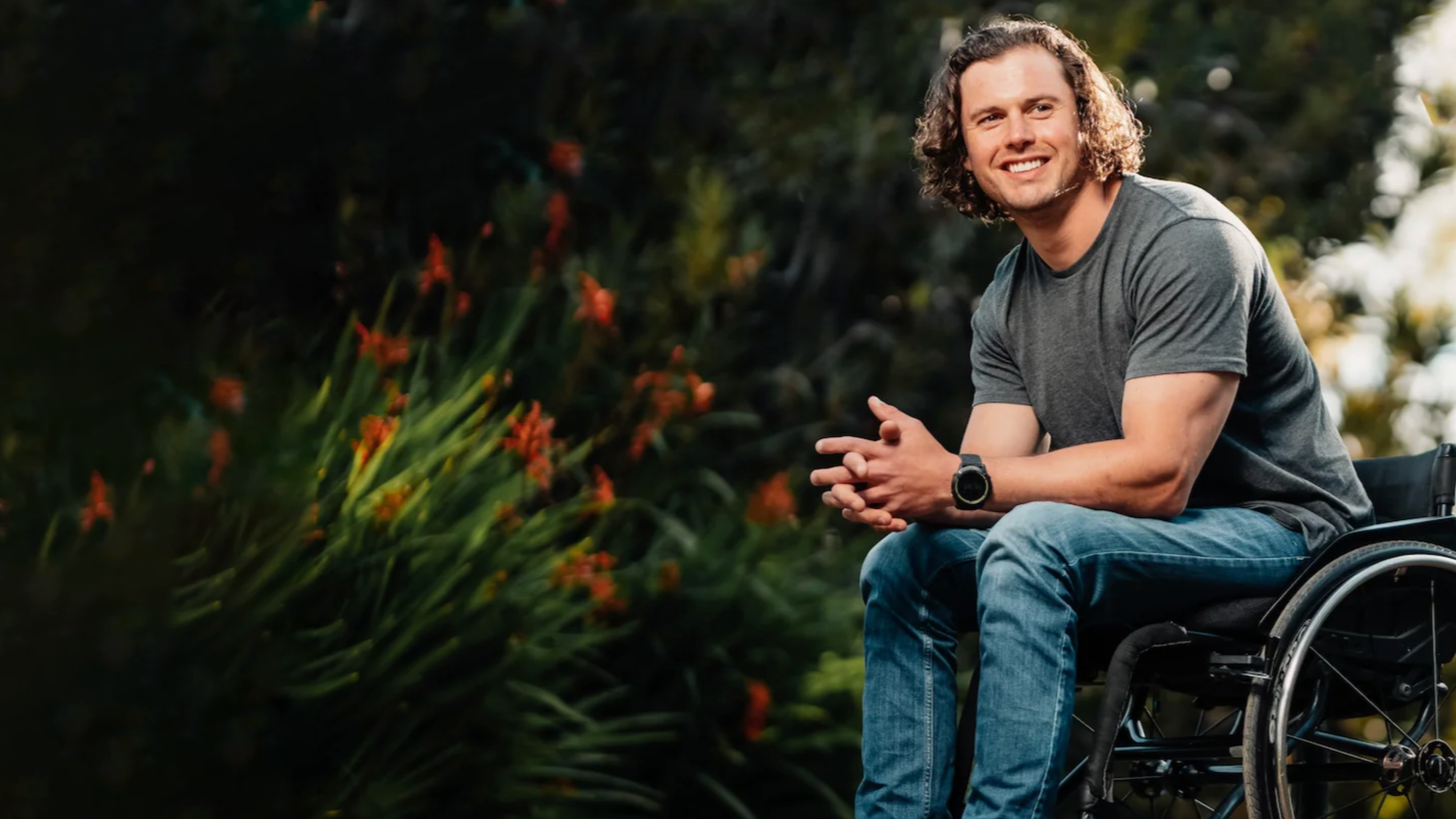 Young man with curly hair sitting in a wheelchair outdoors, smiling and looking at the camera, with greenery and orange flowers in the background.