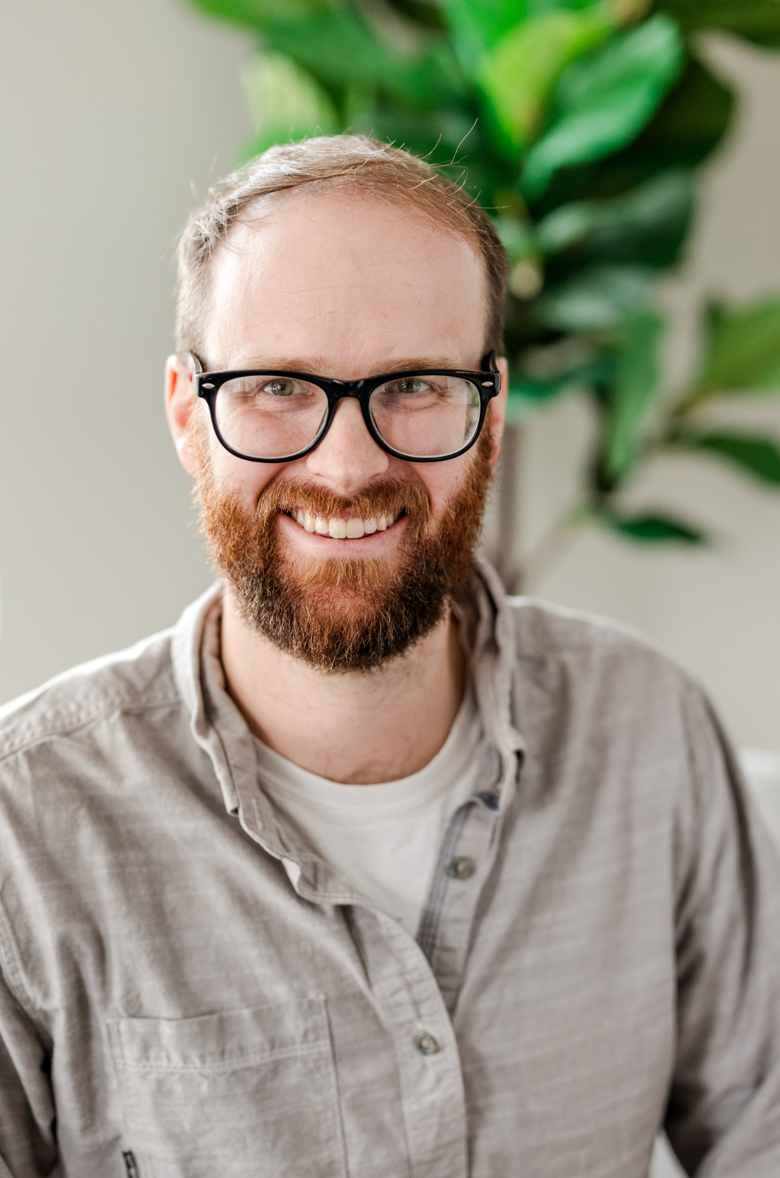 A smiling man with glasses, a beard, and short hair, wearing a light-colored casual shirt, sitting indoors with a potted plant in the background.