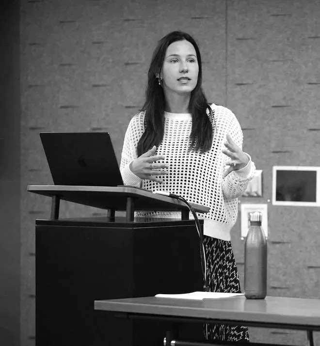 Elise Maddox, a woman with long dark hair standing next to a laptop on a lectern, speaking and gesturing with her hands in a presentation or lecture setting. She is wearing a white knit sweater. There is a water bottle on a table in front of her.
