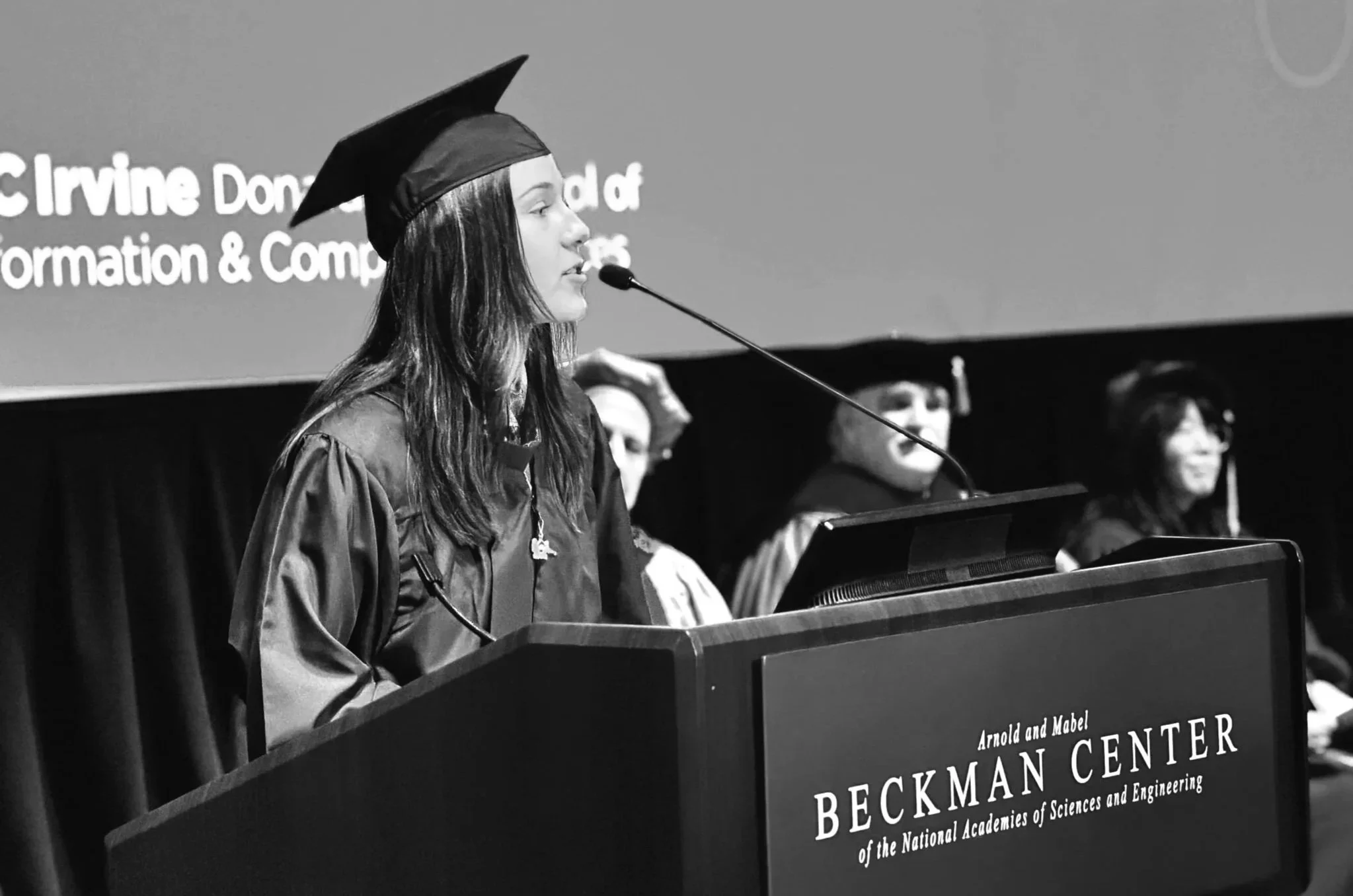 Elise Maddox in academic regalia giving a speech at a podium during a graduation ceremony at the Beckman Center of the National Academies of Sciences and Engineering.