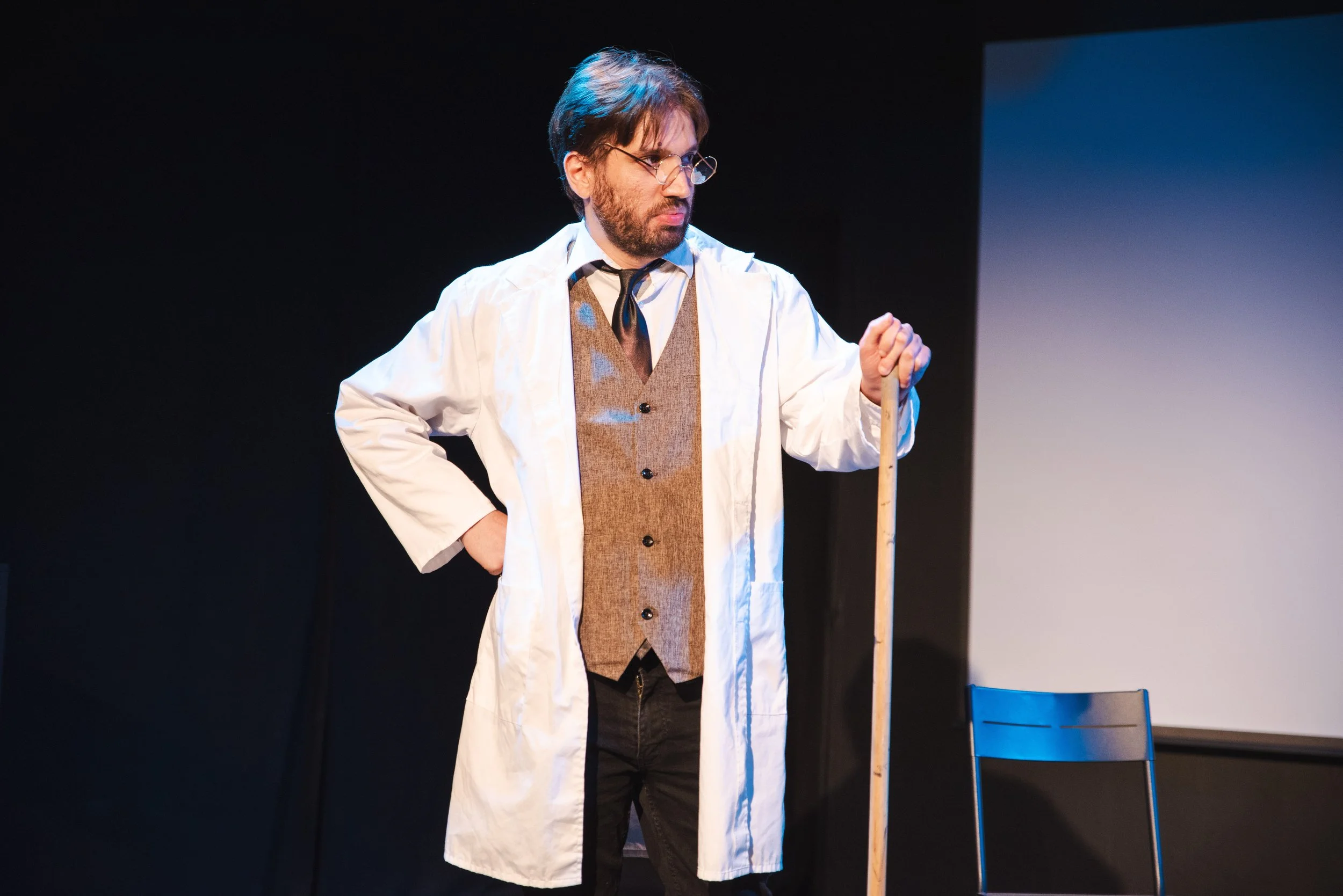 Man wearing glasses, a white lab coat, and a brown vest, holding a wooden stick, standing on a stage with a black background and a chair nearby.