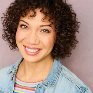 Close-up of a woman with curly hair smiling, wearing a denim jacket and a colorful striped shirt, against a light pink background.
