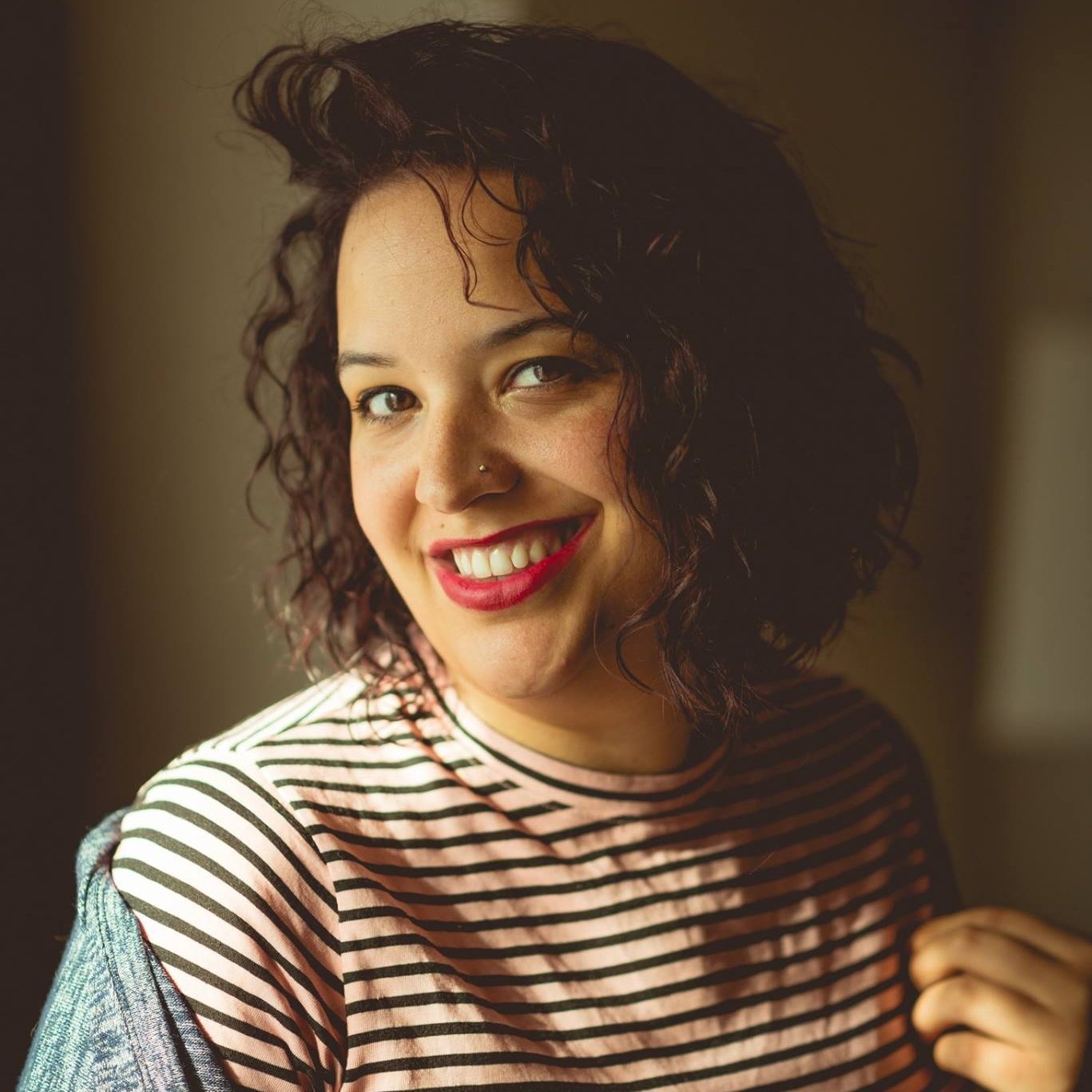 A woman with curly dark hair, red lipstick, and a nose piercing smiling at the camera. She is wearing a striped shirt and has a jacket over her shoulder, standing indoors.