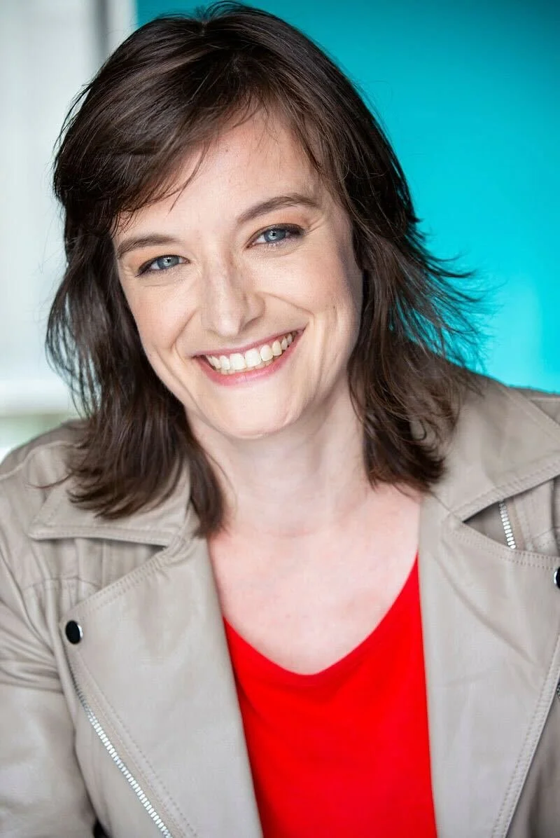 A woman with brown hair and blue eyes smiling at the camera, wearing a red top and a beige jacket, posed against a bright, colorful background.