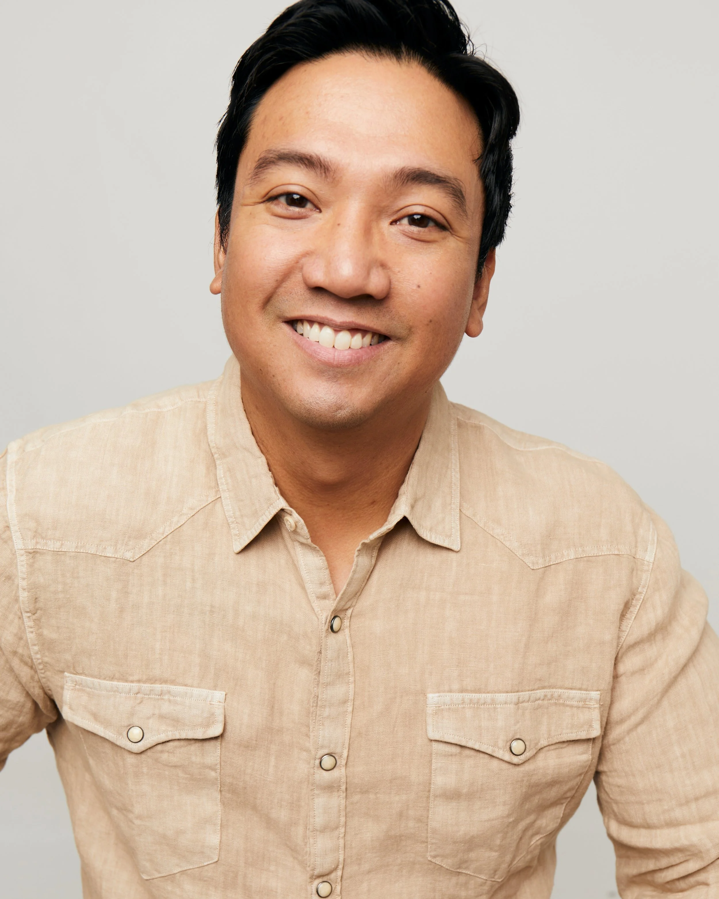 A smiling man with black hair wearing a beige button-up shirt against a plain light gray background.