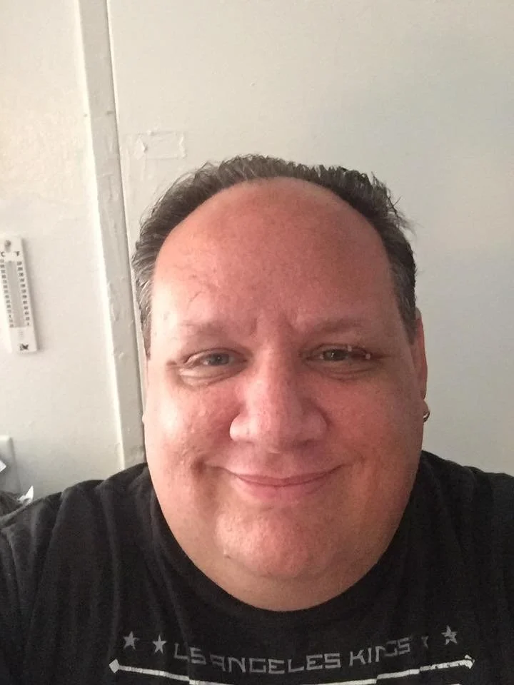 Close-up photo of a smiling man with short dark hair, wearing a black T-shirt, indoors with a plain wall and thermostat in the background.