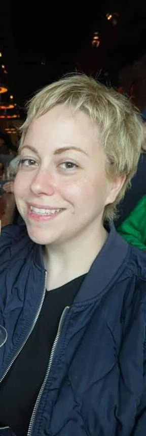 A woman with short blonde hair smiling at a restaurant or bar with warm lighting.