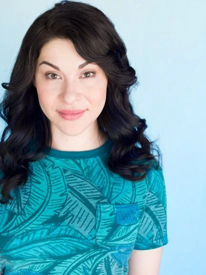Portrait of a woman with long, dark, wavy hair wearing a blue tropical-patterned shirt, standing against a light blue background.