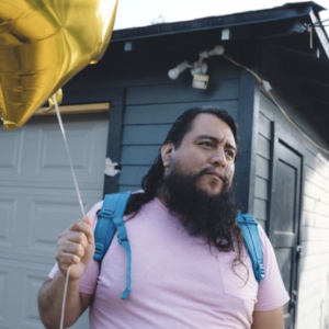 A man with long dark hair and beard, wearing a pink T-shirt and blue backpack, holding a gold balloon, standing outside a dark blue house.