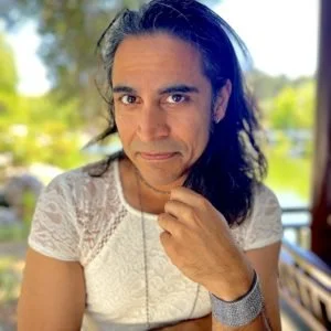 A woman with long dark hair and tan skin, wearing a white lace top, sitting outdoors with trees and a lake in the background.