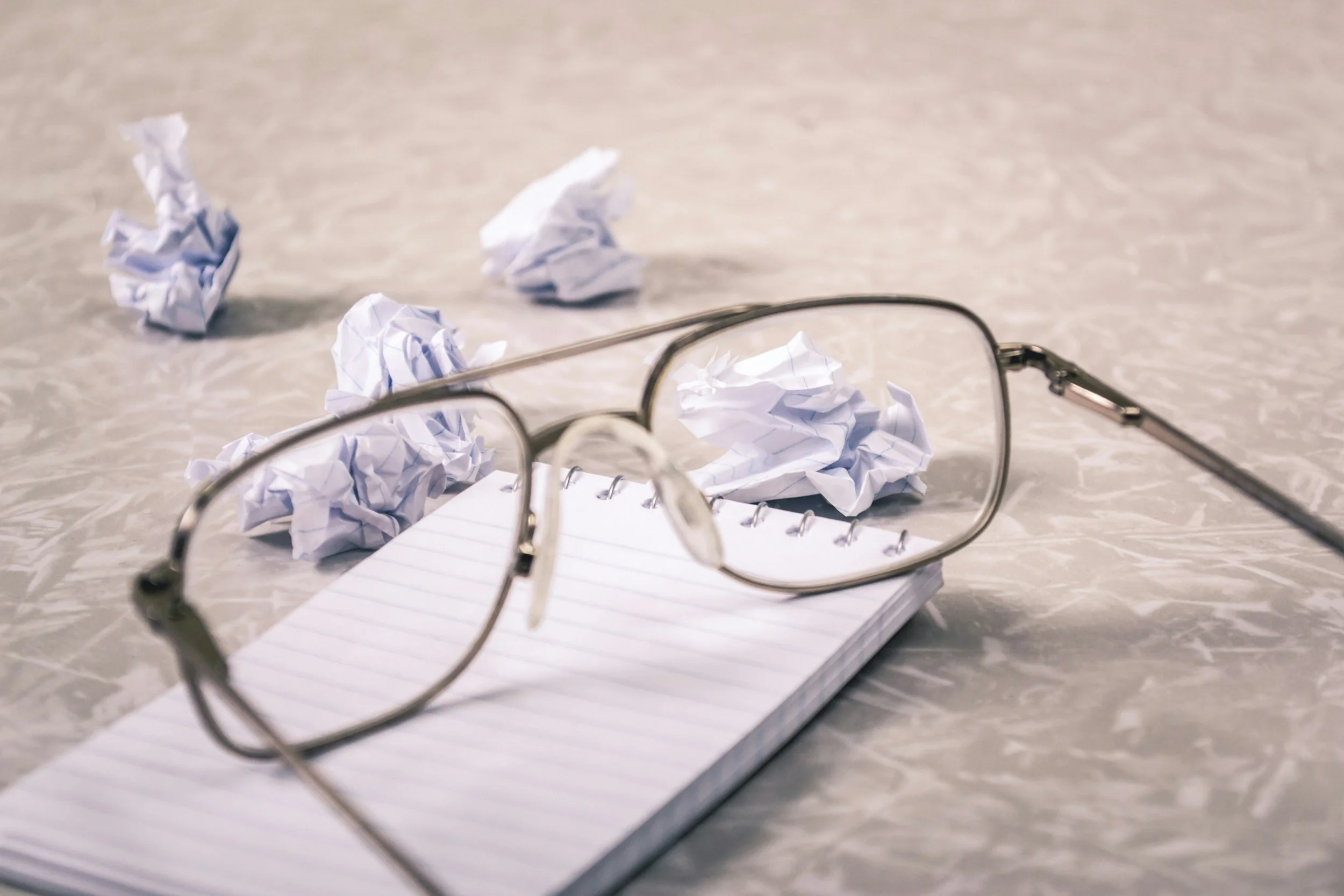 A pair of eyeglasses resting on an open notebook, surrounded by crumpled pieces of paper on a gray surface.