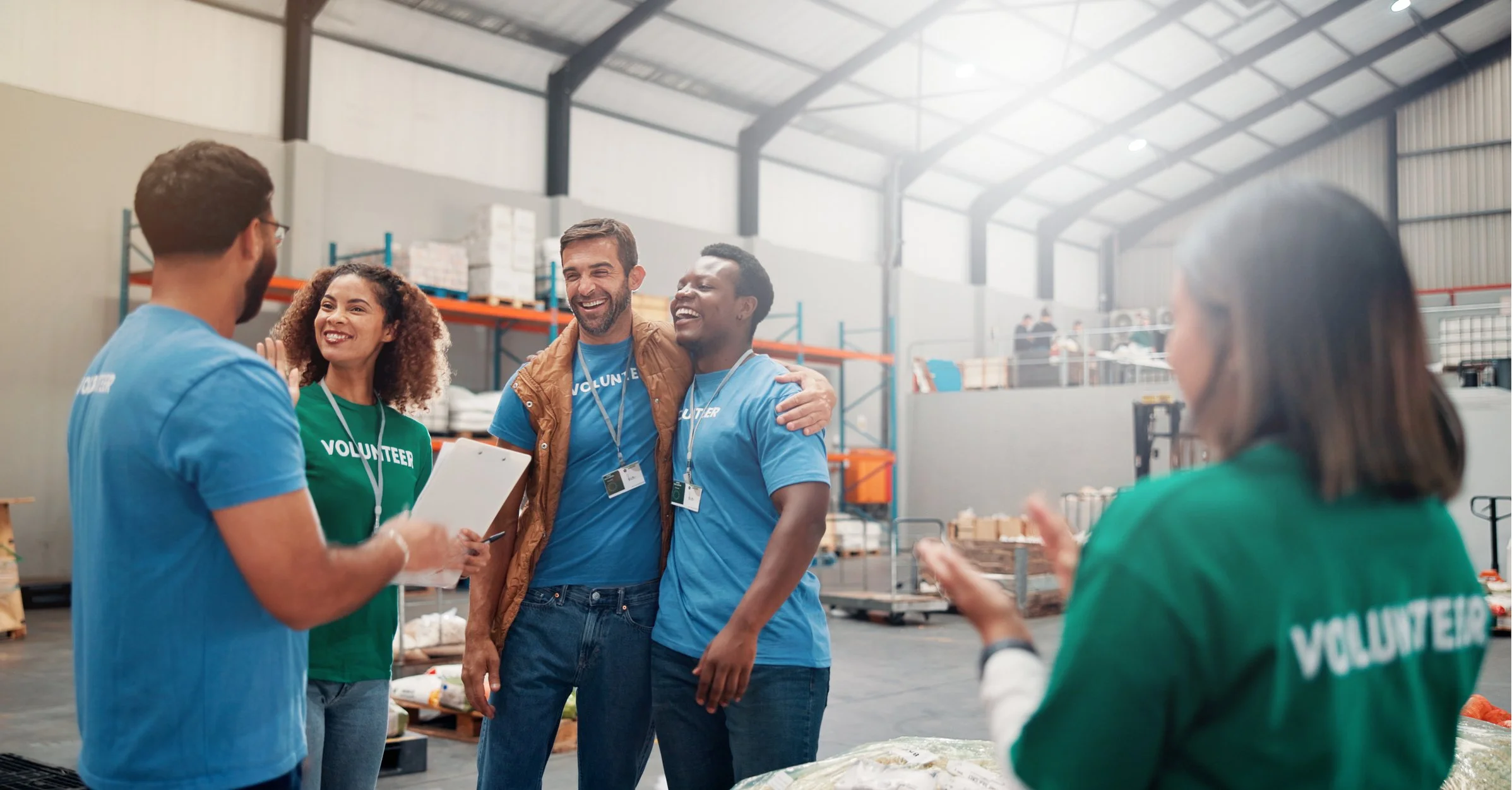 Group of volunteers smiling and talking inside a warehouse, some wearing blue and green volunteer shirts, surrounded by shelves and supplies.