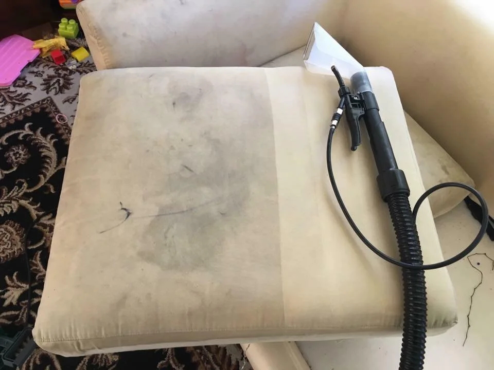 A beige upholstered chair with visible stains and marks, with a black cleaning or blowing tool resting on it. The surrounding area includes some children's toys and a patterned carpet.