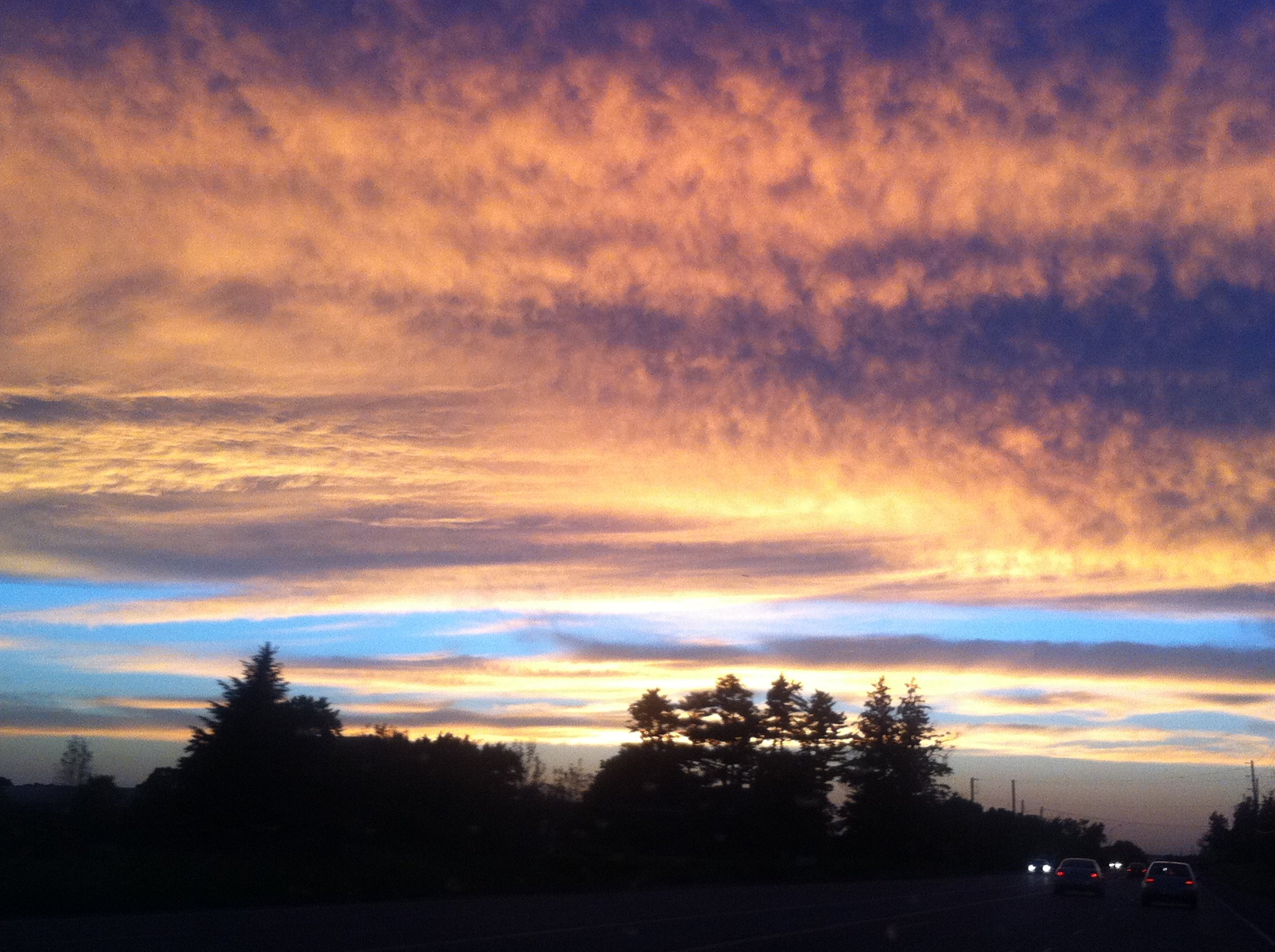 A colorful sunset sky with orange, pink, purple, and blue clouds over a silhouette of trees and a highway with cars.